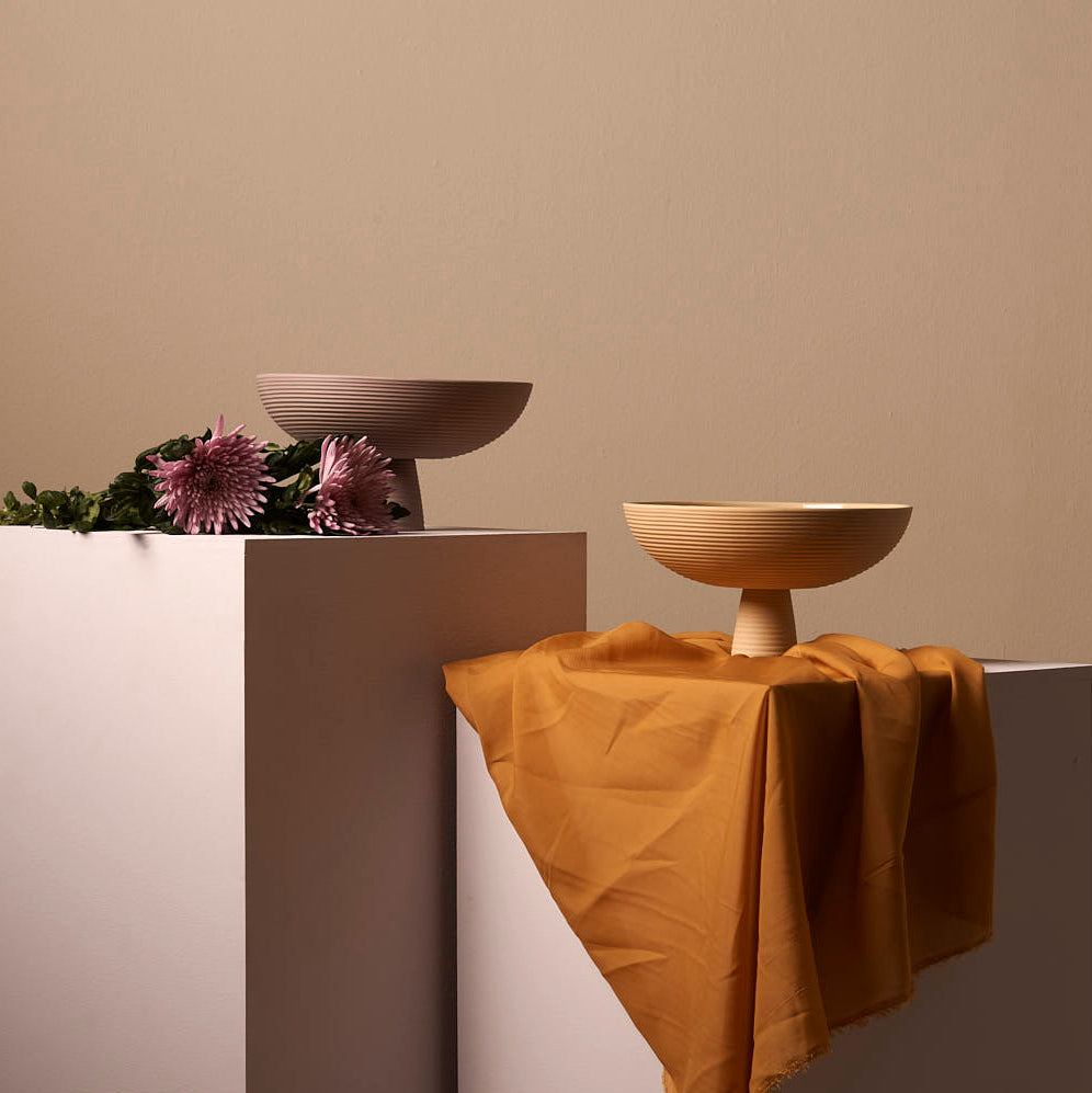 Two ceramic bowls from Schneid, including the Dais, are elegantly presented on light pedestals. One sits on a mustard cloth, while the Dais bowl showcases pink flowers and green foliage. A soft beige wall completes this charming setup for modern homes.