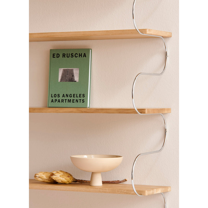 Wooden shelves with a silver wavy bracket hold the green book Ed Ruscha: Los Angeles Apartments and a ceramic Dais bowl by Schneid. Two dried artichokes beside the bowl enhance the charm of modern homes.