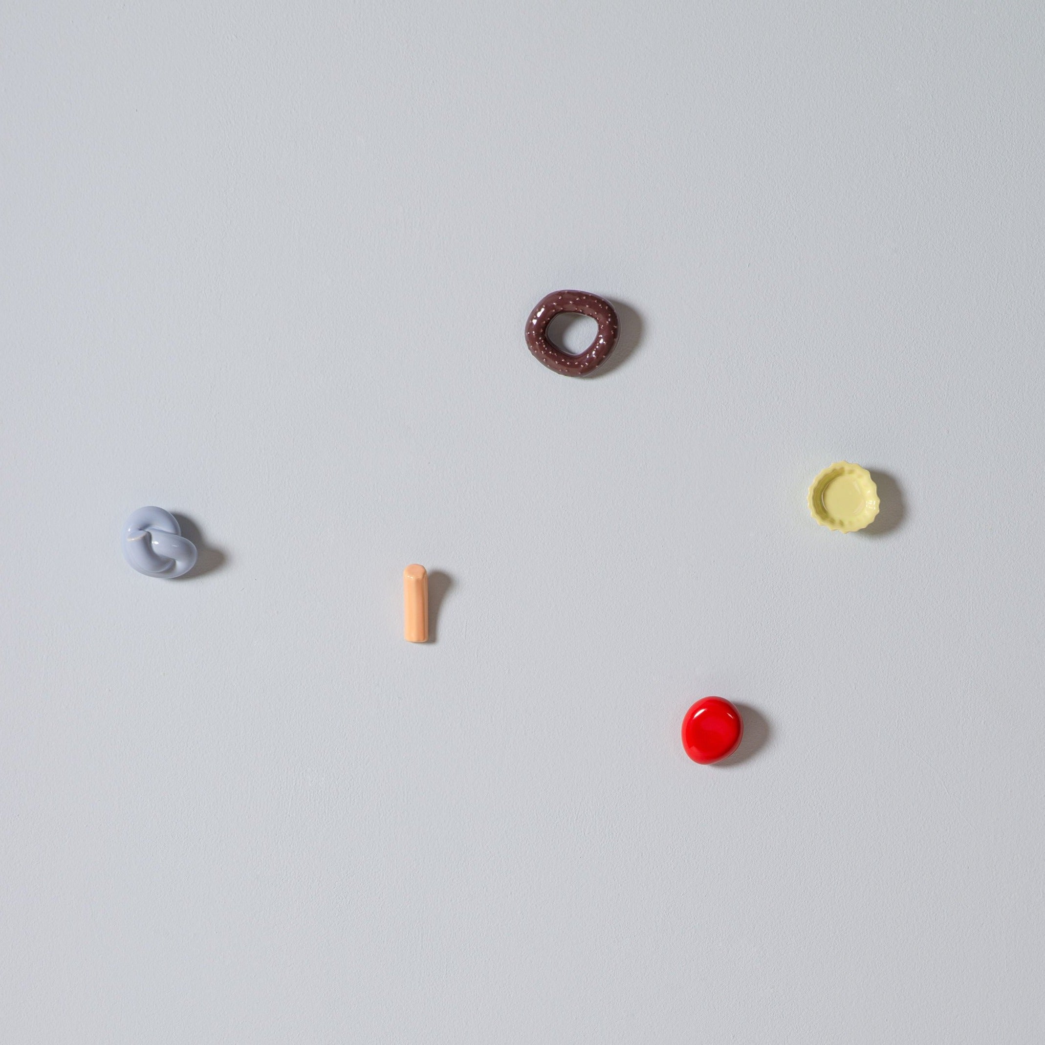 Schneids Fossil Hooks artfully features vibrant organic shapes on a gray backdrop, with a brown loop, yellow ripple, red circle, beige cylinder, and blue twist. The design echoes abstract colorful glazes splashed across the canvas.