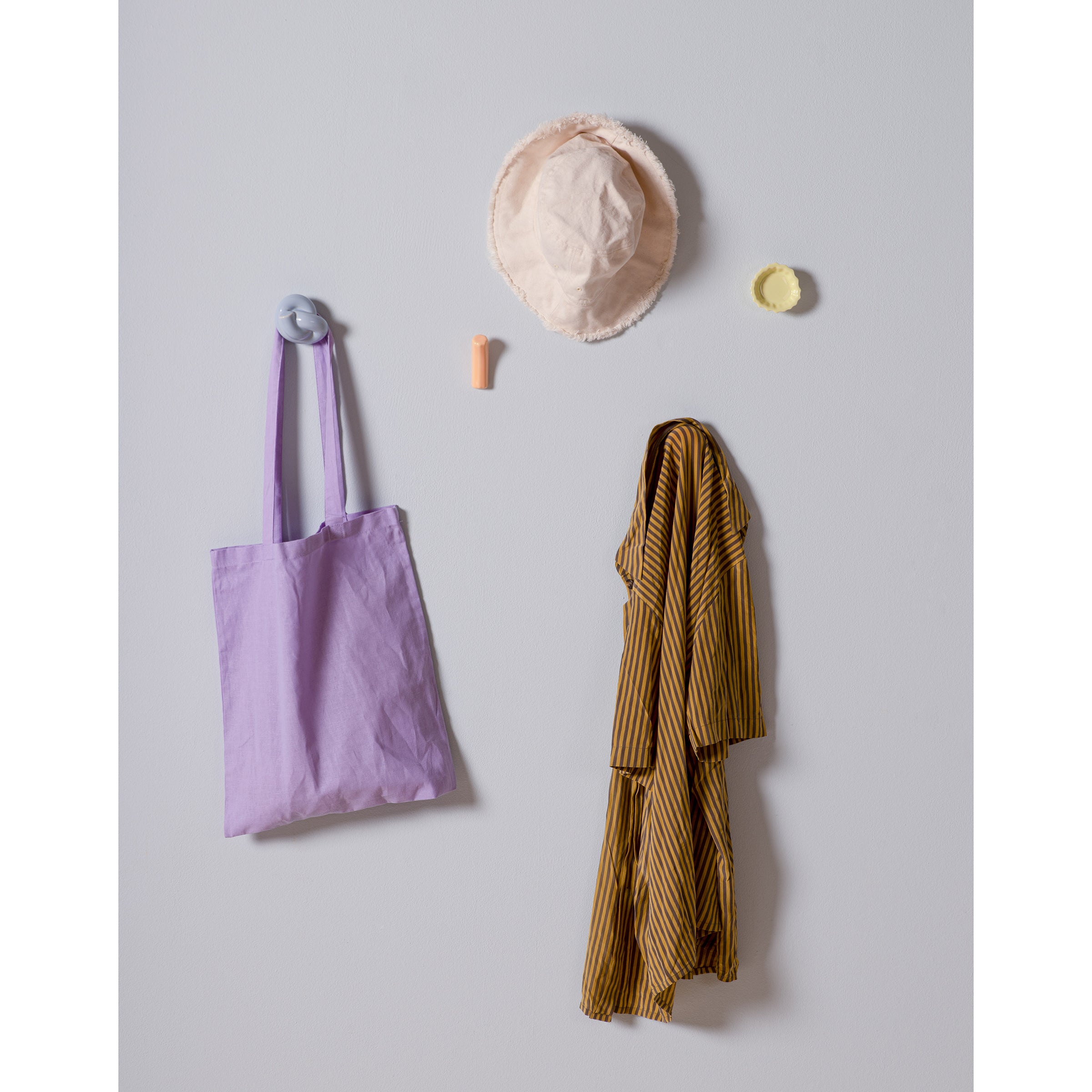 A minimalistic wall setup includes a lavender tote and mustard-striped shirt on Schneid Fossil Hooks. Above, a pink bucket hat, small orange tube, and round yellow item with colorful glazes add charm to the plain, light-colored backdrop.