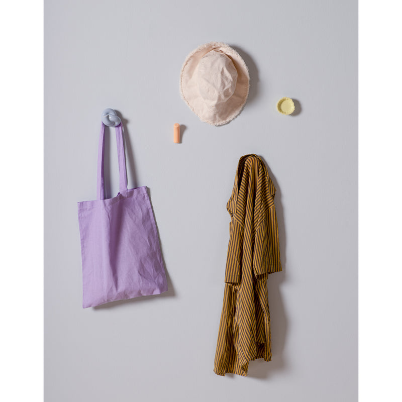 A minimalistic wall setup includes a lavender tote and mustard-striped shirt on Schneid Fossil Hooks. Above, a pink bucket hat, small orange tube, and round yellow item with colorful glazes add charm to the plain, light-colored backdrop.