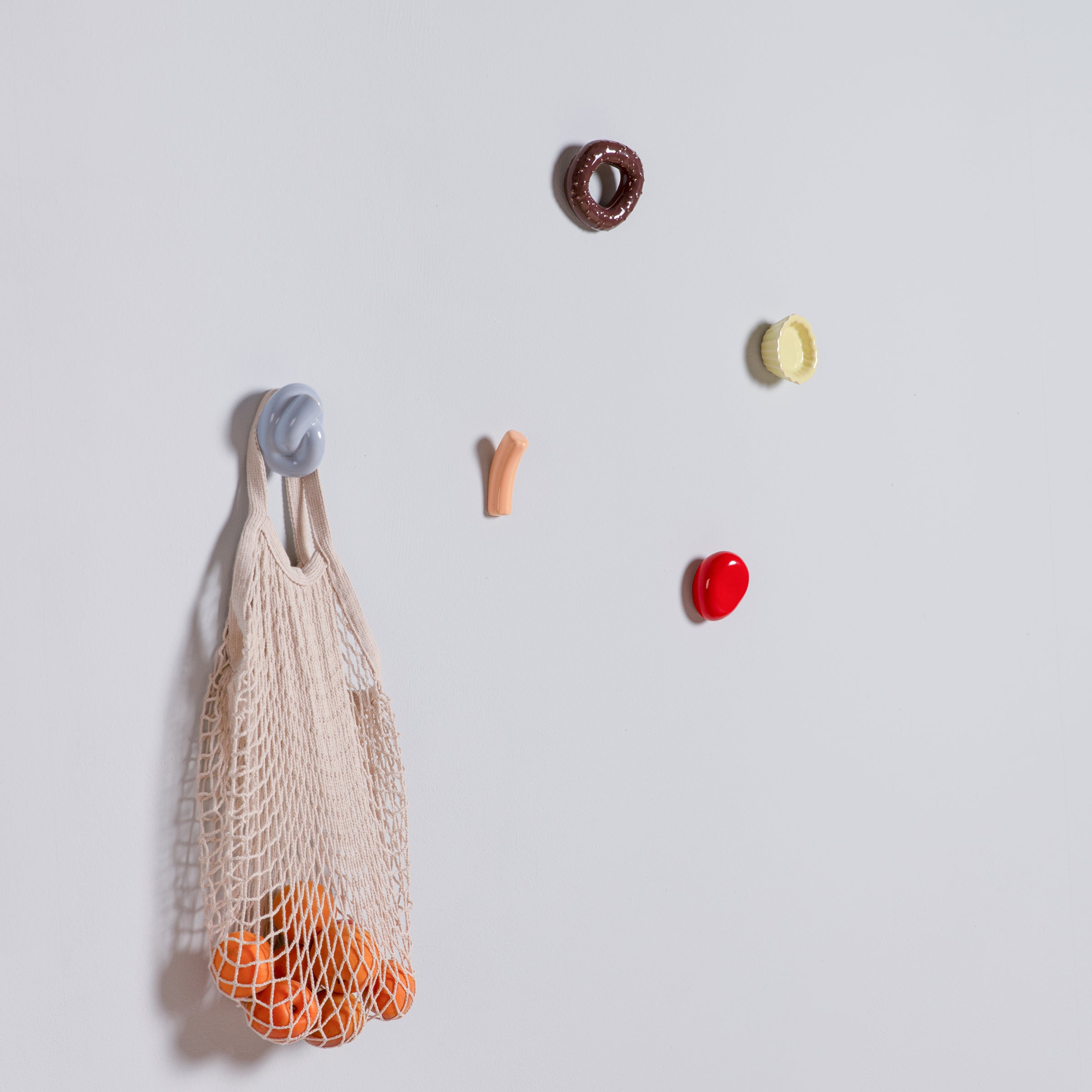 A net bag with oranges hangs on Schneids Fossil Hooks against a white backdrop. These hooks have vibrant glazes and organic shapes, showcasing colors like blue, brown, pink, red, and yellow.