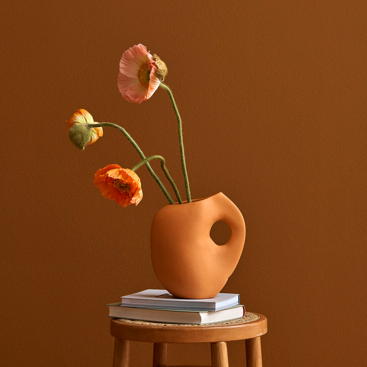 A minimalist scene presents a Schneid Aura I vase with a circular handle, holding bright orange and pink poppies. This stoneware piece rests on a few books atop a wooden stool against a warm brown backdrop.