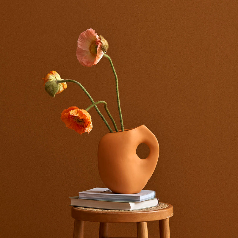 A minimalist scene presents a Schneid Aura I vase with a circular handle, holding bright orange and pink poppies. This stoneware piece rests on a few books atop a wooden stool against a warm brown backdrop.