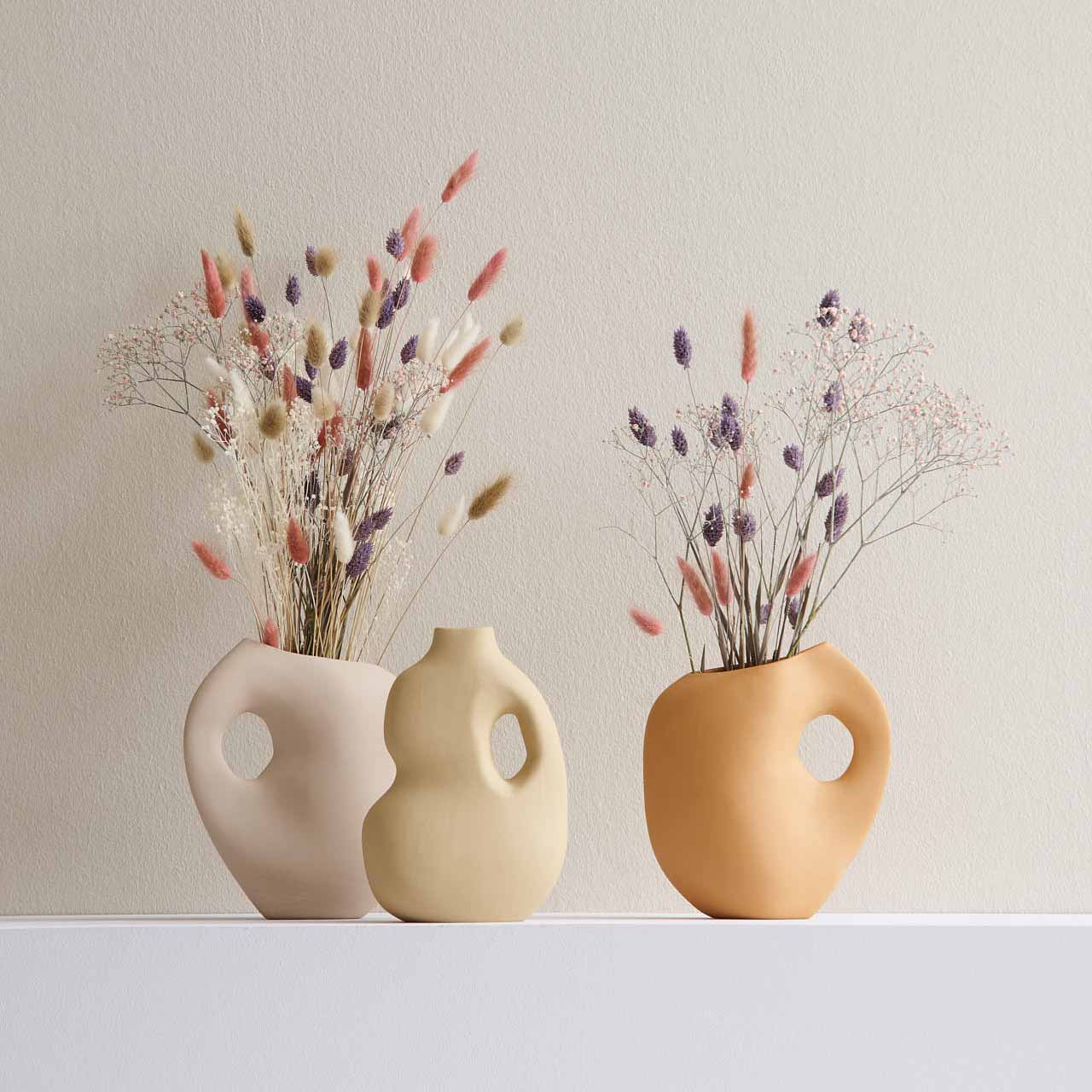 Two uniquely shaped Aura vases with handles rest on a white surface. One, a beige stoneware object with a dry floral arrangement; the other, mustard with similar flowers. The neutral-toned wall backdrop creates an elegant and minimalist scene.
