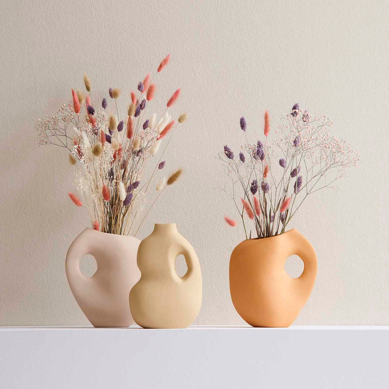 Three uniquely shaped Schneid Aura II vases with large holes and pastel shades are displayed against a light background. These stoneware objects hold dried flowers in pink, purple, and cream, creating a soft, artistic composition.