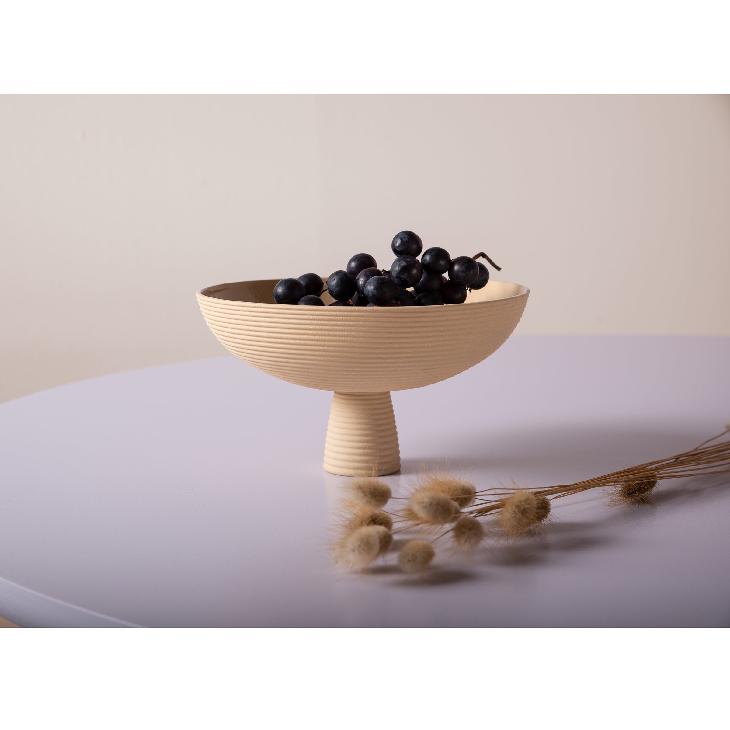 Schneids Dais, a beige ribbed ceramic pedestal bowl, cradles dark grapes atop a light purple surface. Dried grass stalks lie in front, bringing a natural touch to modern homes with a soft cream background.