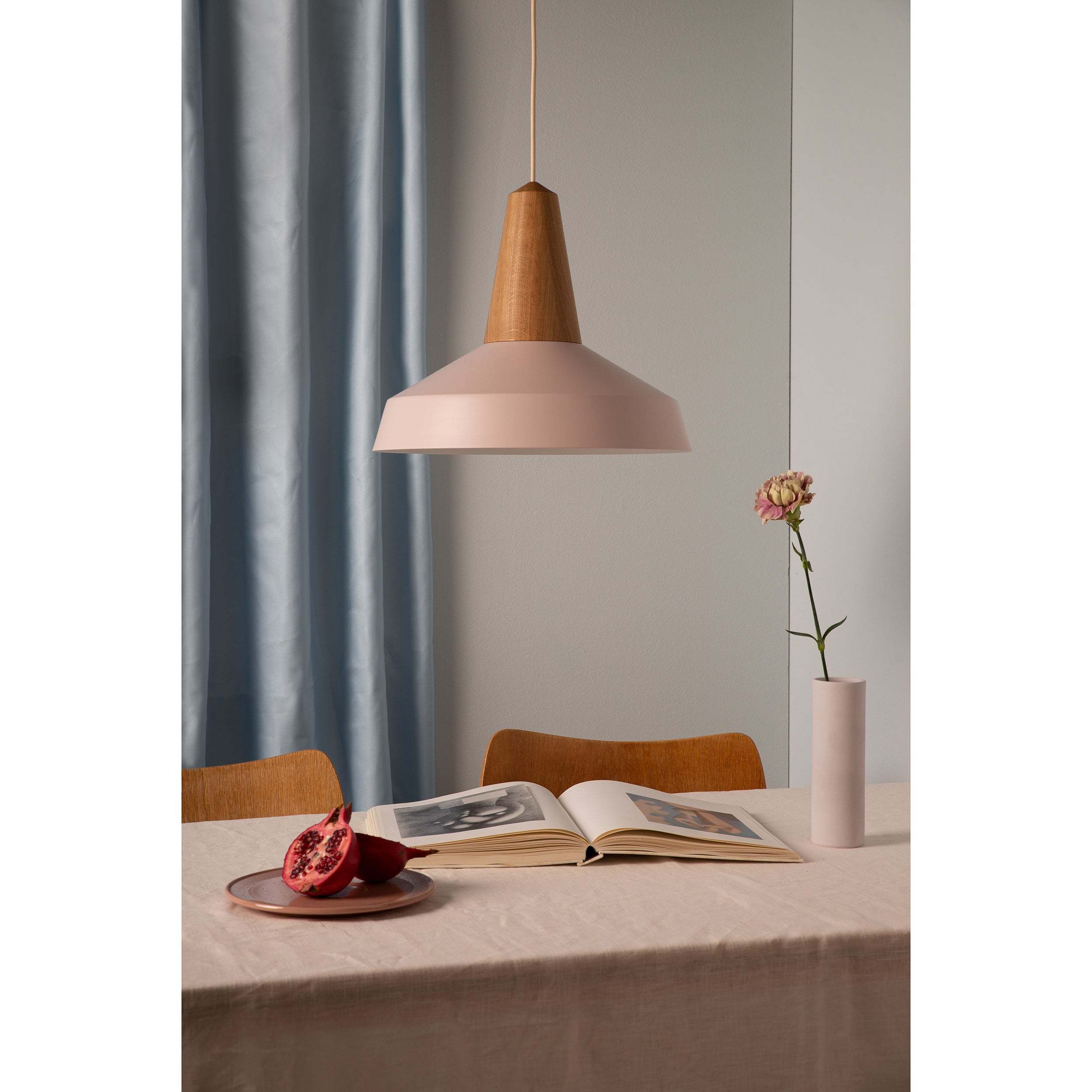 A pale rose Eikon Circus pendant lamp by Schneid illuminates a table with a white cloth. On the table, theres an open book, half a pomegranate on a pink plate, and a flower in a tall vase. A light blue curtain softly frames this modern scene in the background.