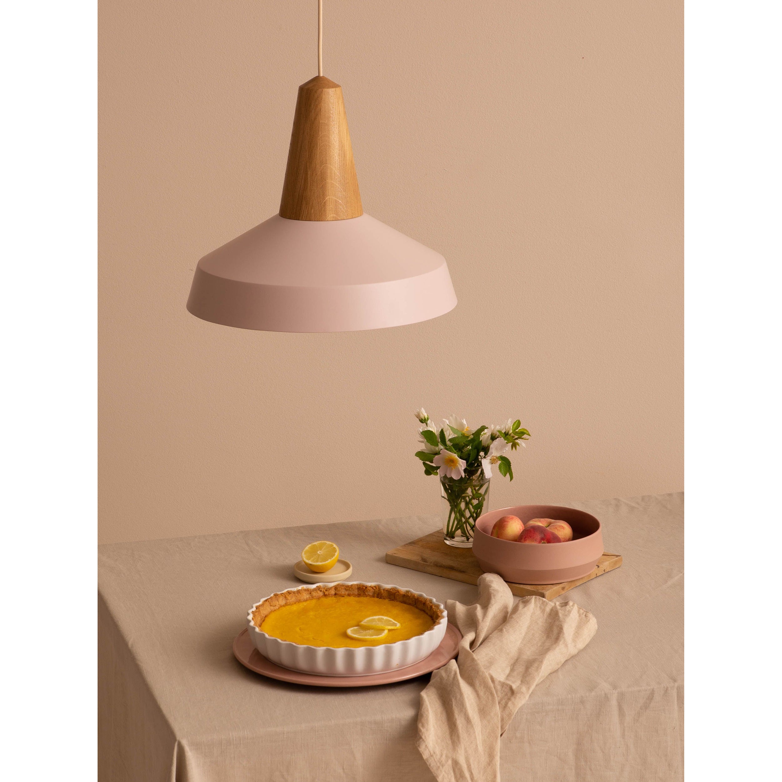 A minimalist table setting boasts a lemon tart on a plate, peaches in a bowl, and white flowers in a vase on a beige tablecloth. An Eikon Circus Pale Rose light by Schneid with a wooden top adds contemporary flair against the soft, neutral beige wall.