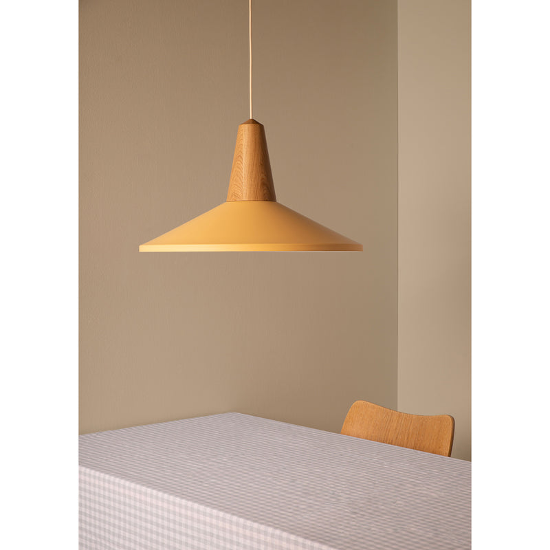A minimalist room showcases a wooden chair from certified wood and a table with a checkered cloth. Above, the Eikon Shell Olive by Schneid, a modern cone-shaped pendant light on sale, casts a warm yellow glow against the beige wall.