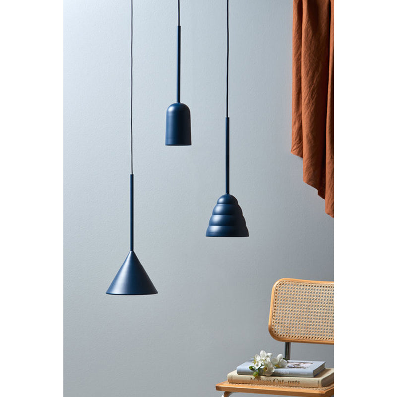 Three Figura Cone pendant lights by Schneid hang from the ceiling against a light wall. Below is a wicker chair with a small stack of books and a white flower. A rust-colored curtain partially enters the frame from the right.