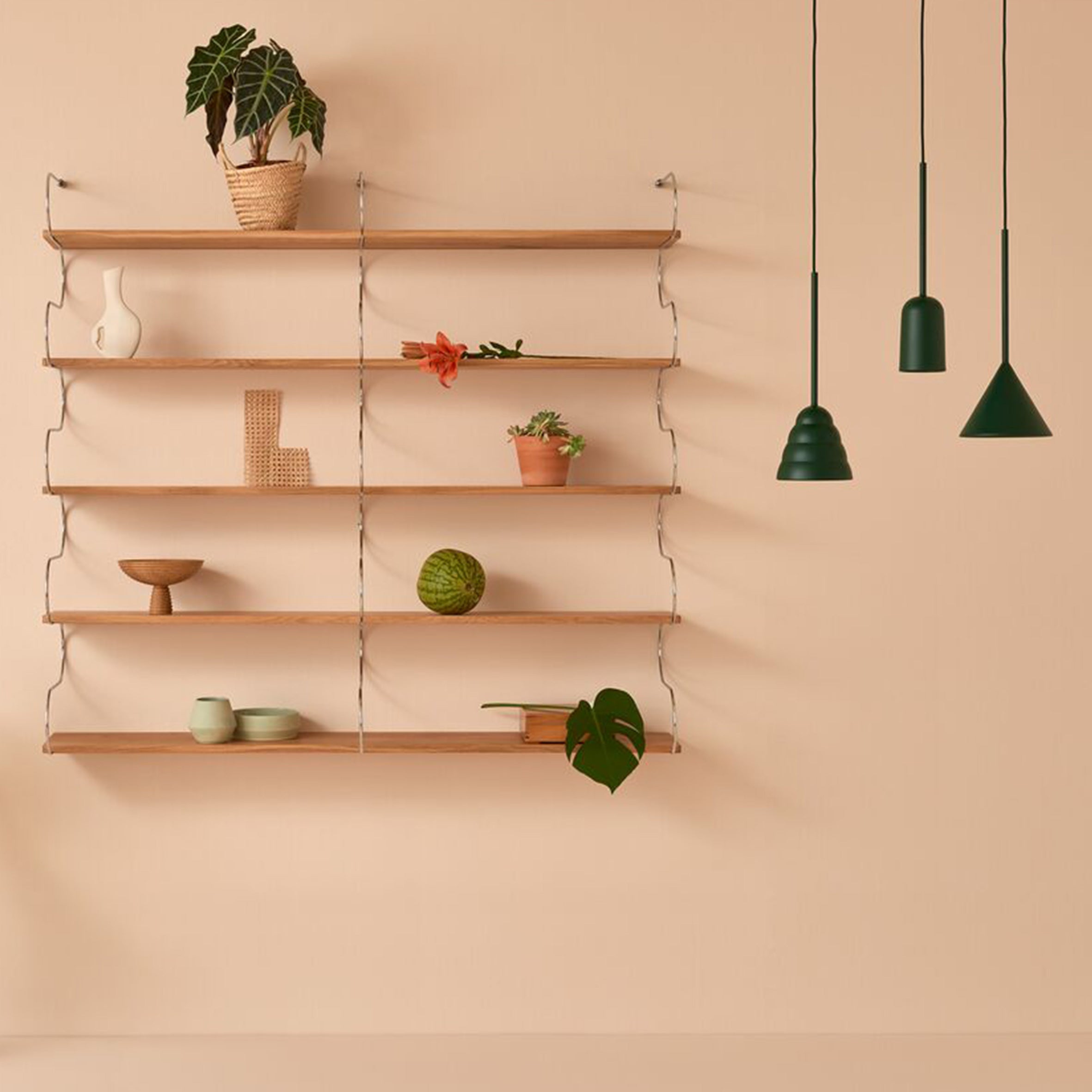 In this minimalist room, light wood shelves mounted on a beige wall hold a small vase with a flower, decorative items, and a potted plant. To the right, three Schneid Figura Cone pendant lights in green create a sculptural silhouette for an elegant modern touch.