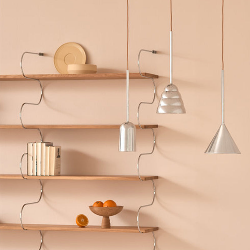 A minimalist room features wooden shelves with books, a plate, and a bowl of oranges. Hanging against the beige wall are three modern pendant lights: an industrial sculptural luminaire, the Schneid Figura Cone - Sale, and a ribbed dome.