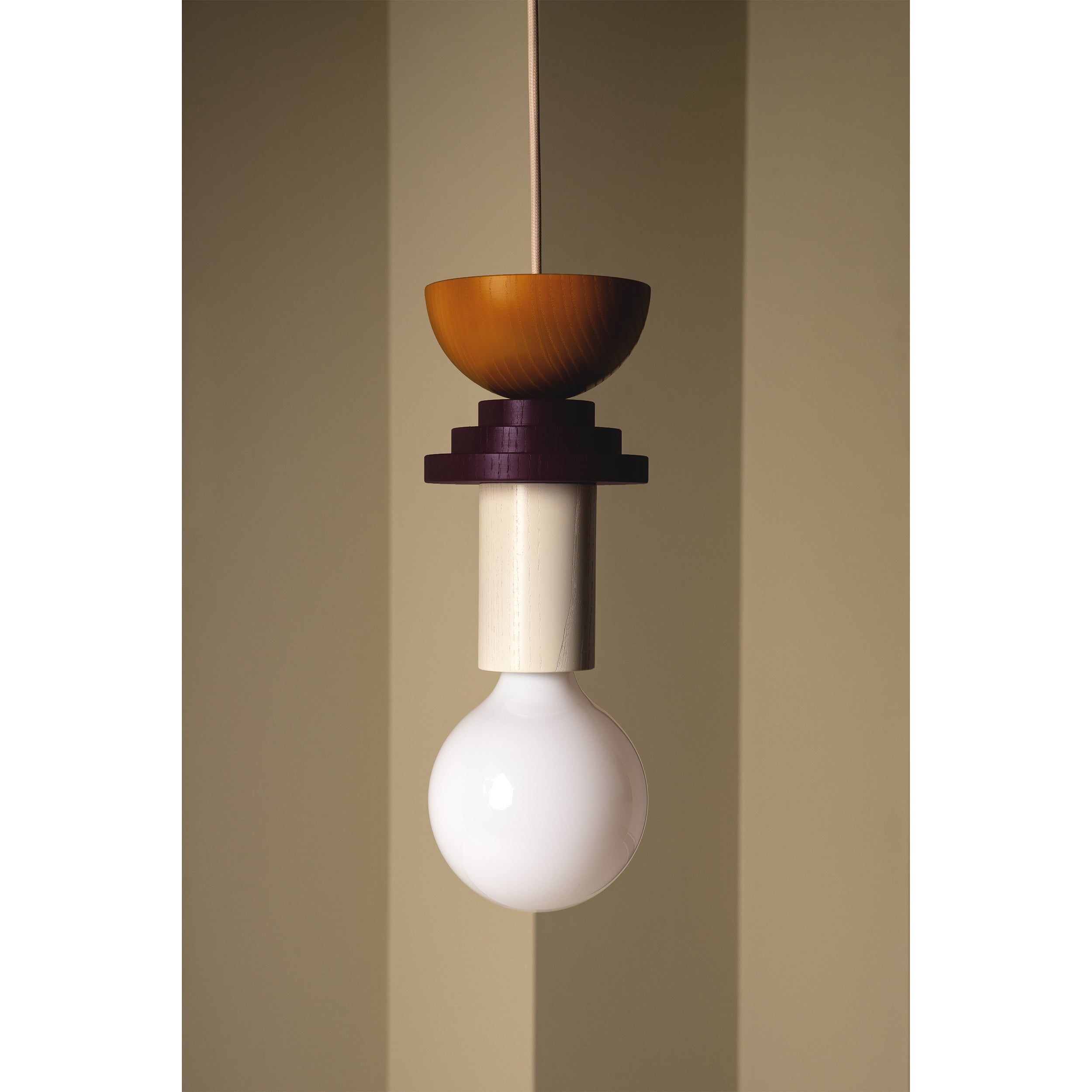 Introducing the Junit Karma by Schneid, a modern modular pendant lamp with a unique design featuring an ash wood cylindrical base, white bulb, round orange cap, and stacked dark rings. It complements walls adorned with vertical stripes in muted green tones beautifully.