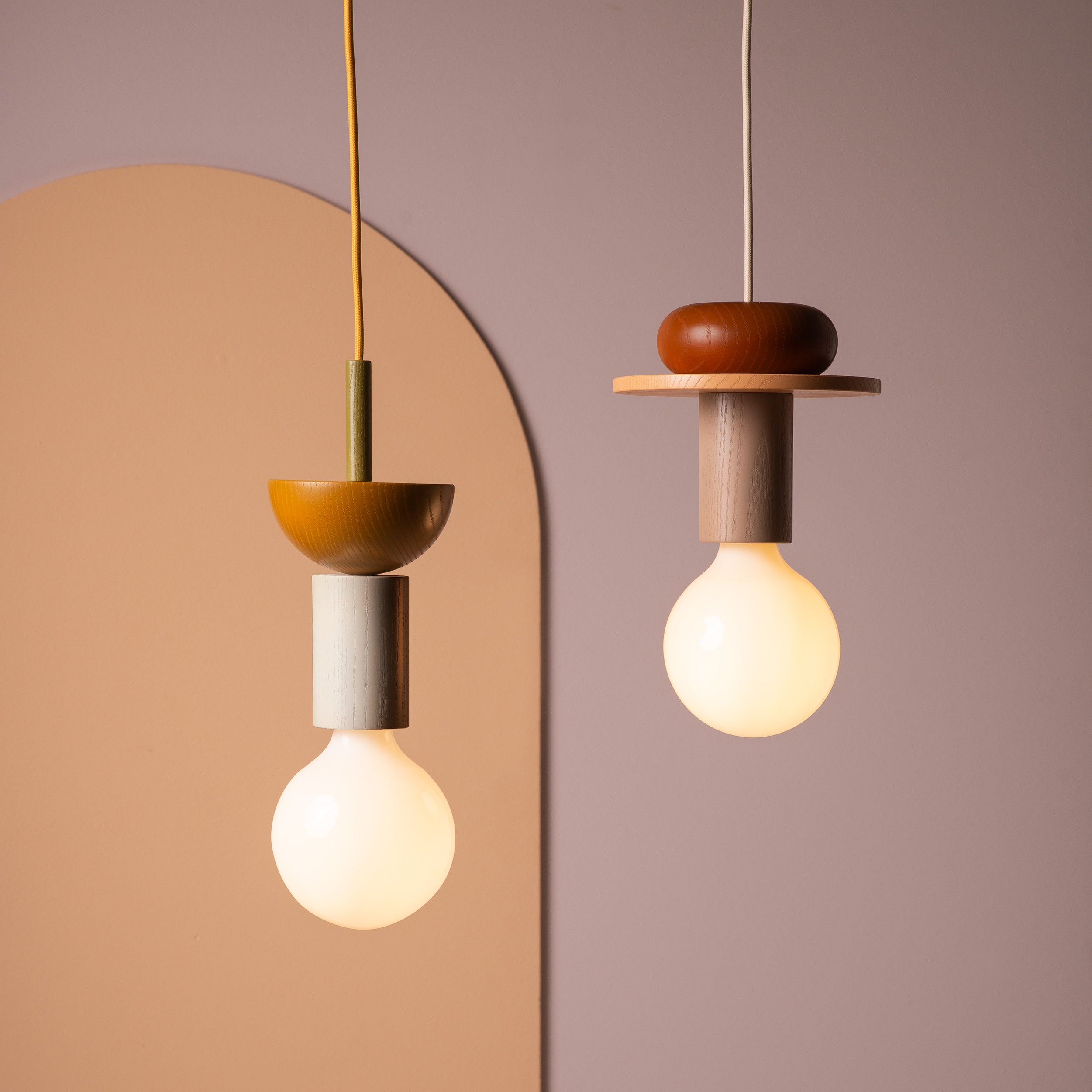 Two modular Junit Pan pendant lamps from Schneid hang against a pastel background. Crafted from geometric ash wood, they emit a soft ambient glow. The left lamp features a gold element, set against an arched panel in muted peach tones.