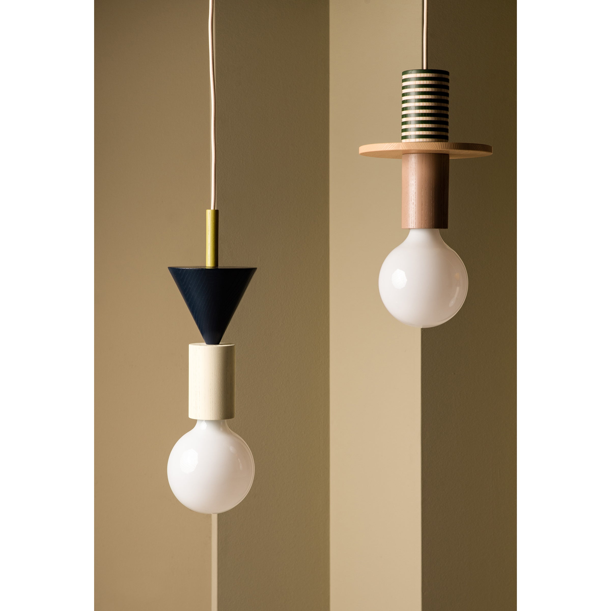 Two modern pendant lights adorn a beige wall. The left, a modular pendant lamp, has a black cone and cylindrical design. The right is the Junit Omen by Schneid, featuring ambient light with striped cap and ash wood accents, both topped with round white bulbs.