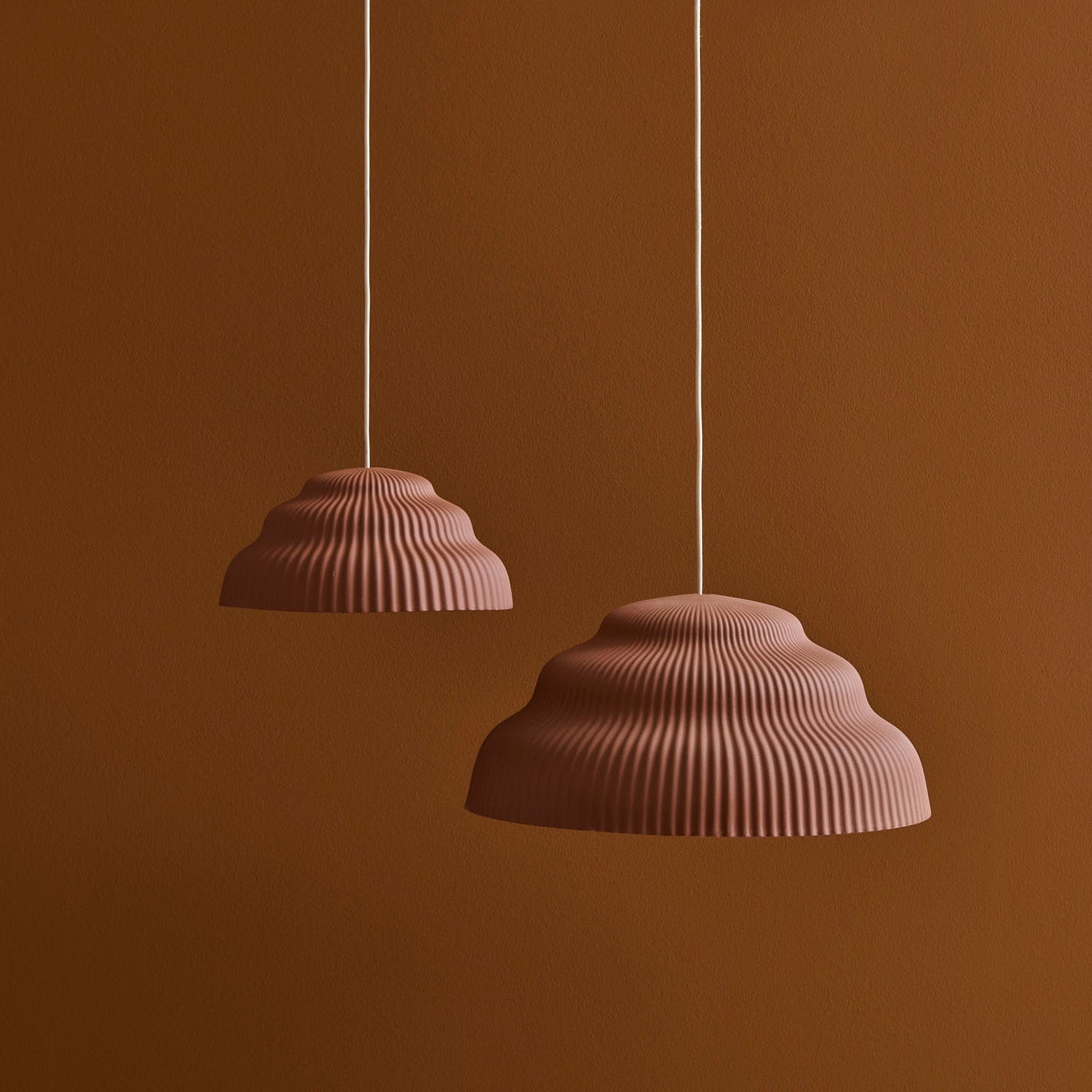 Two Kaskad Rust suspension lamps by Schneid, featuring a ribbed, scalloped design in muted clay color, hang from the ceiling against a warm brown wall. They add an earthy and modern touch to the minimalist setting.