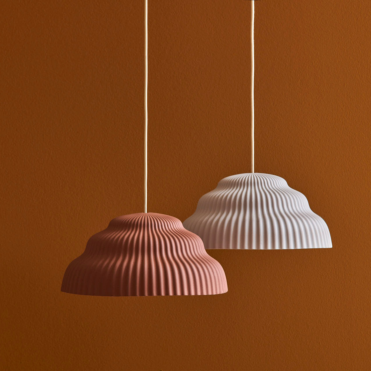 Two Kaskad Rust suspension lamps by Schneid with ribbed, scalloped shades in coral pink and white hang against a terracotta background, suspended by simple white cords.
