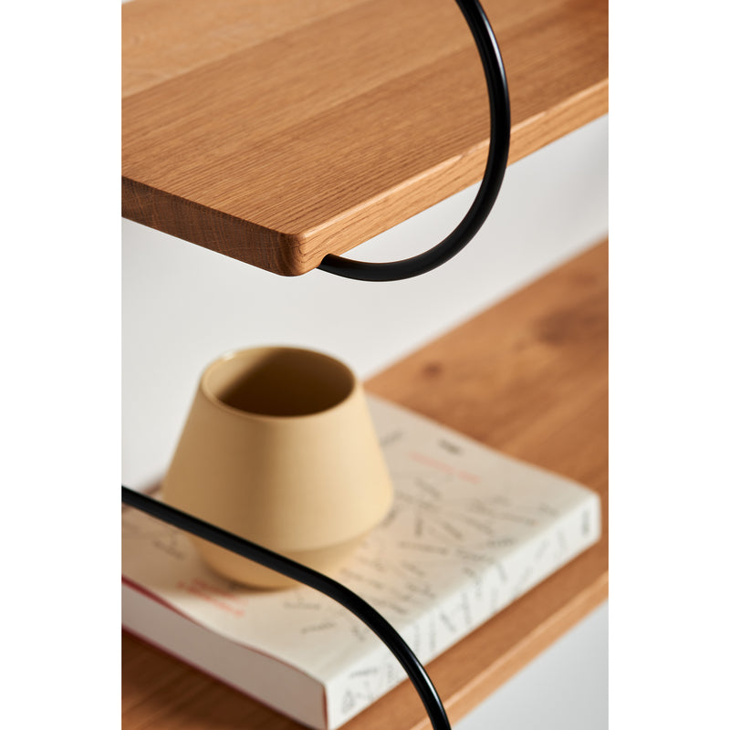A close-up of oak Onda shelves by Schneid featuring a beige cup on a book with a white abstract cover. Nestled within the black metal supports curved lines, it highlights this elegant modular shelving system.