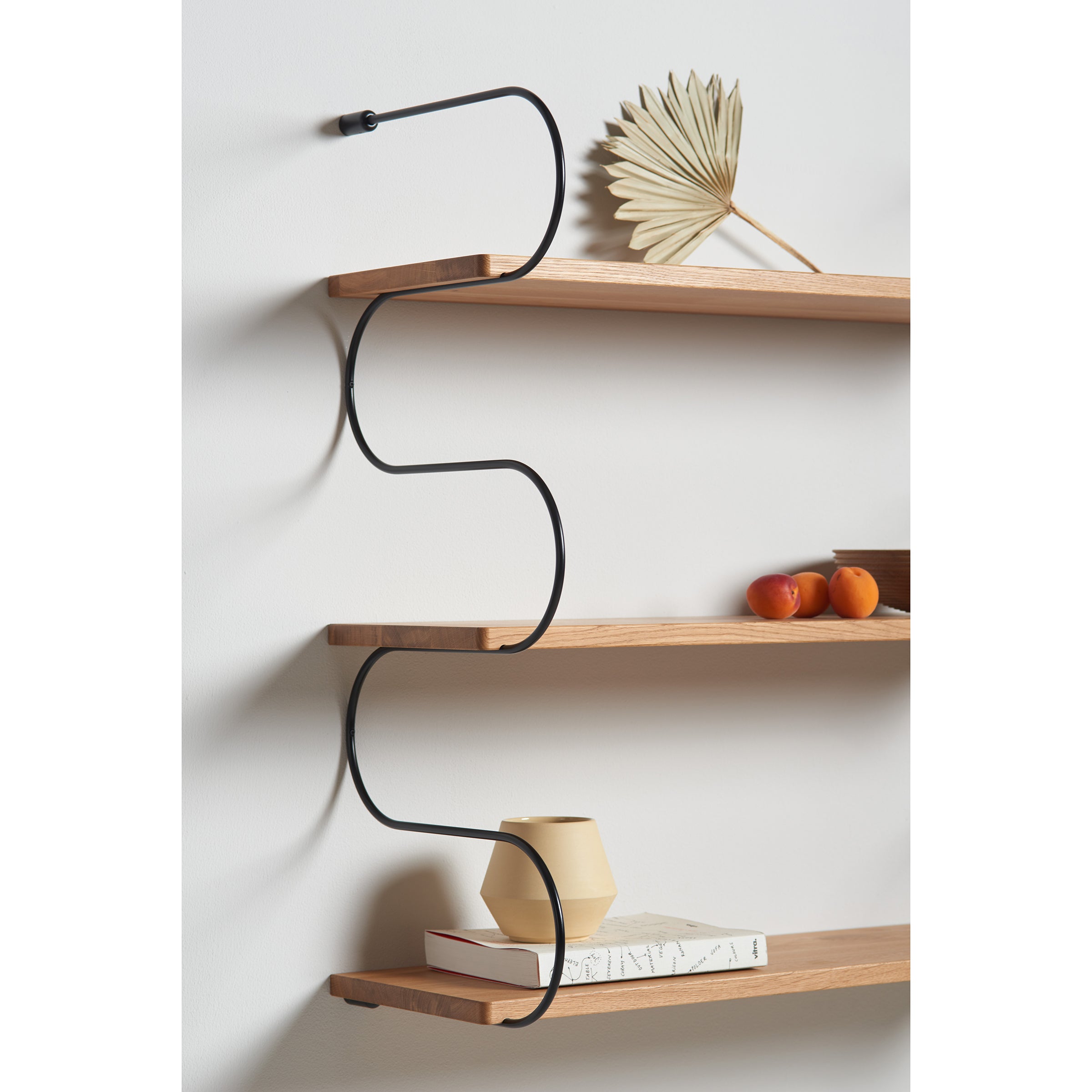 The Onda shelves by Schneid feature oak wood mounted on a wall with black metal zigzag brackets. A decorative dried leaf, oranges, and a beige vase enhance the display, while a book on the bottom shelf completes this stylish modular shelving system.