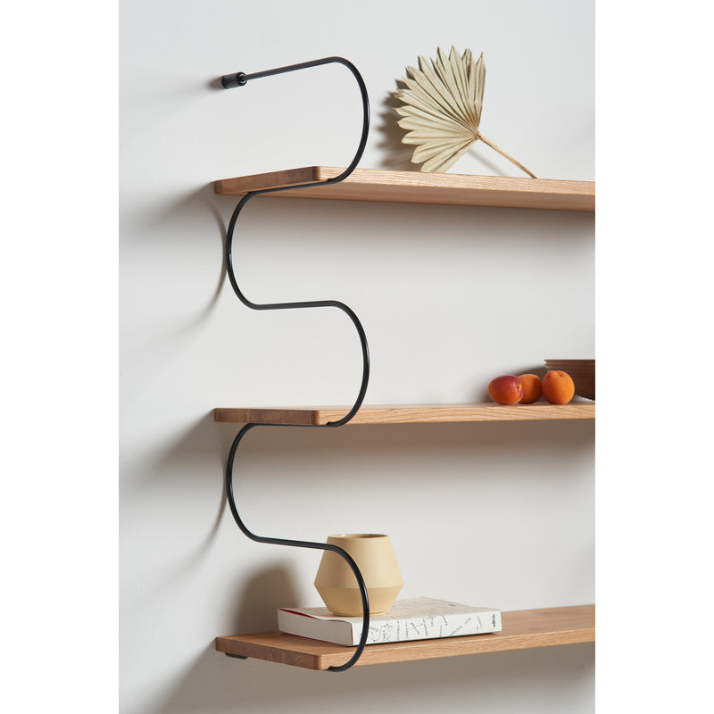 The Onda shelves by Schneid feature oak wood mounted on a wall with black metal zigzag brackets. A decorative dried leaf, oranges, and a beige vase enhance the display, while a book on the bottom shelf completes this stylish modular shelving system.