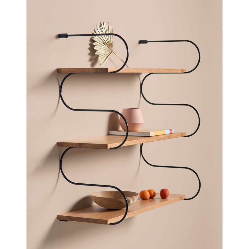 The Onda by Schneid, a modern wall-mounted shelf, boasts a unique black metal frame with curved lines. Its oak wood shelves elegantly display a decorative leaf, pink vase, book stack, bowl, apple, and orange against a beige backdrop.