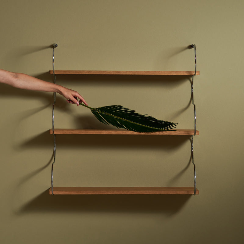 The Onda shelf by Schneid, crafted from minimalist oak wood, features three tiers and hangs on a muted green wall. A hand places a large green leaf on the middle shelf, with the understated background enhancing its sleek curved design.