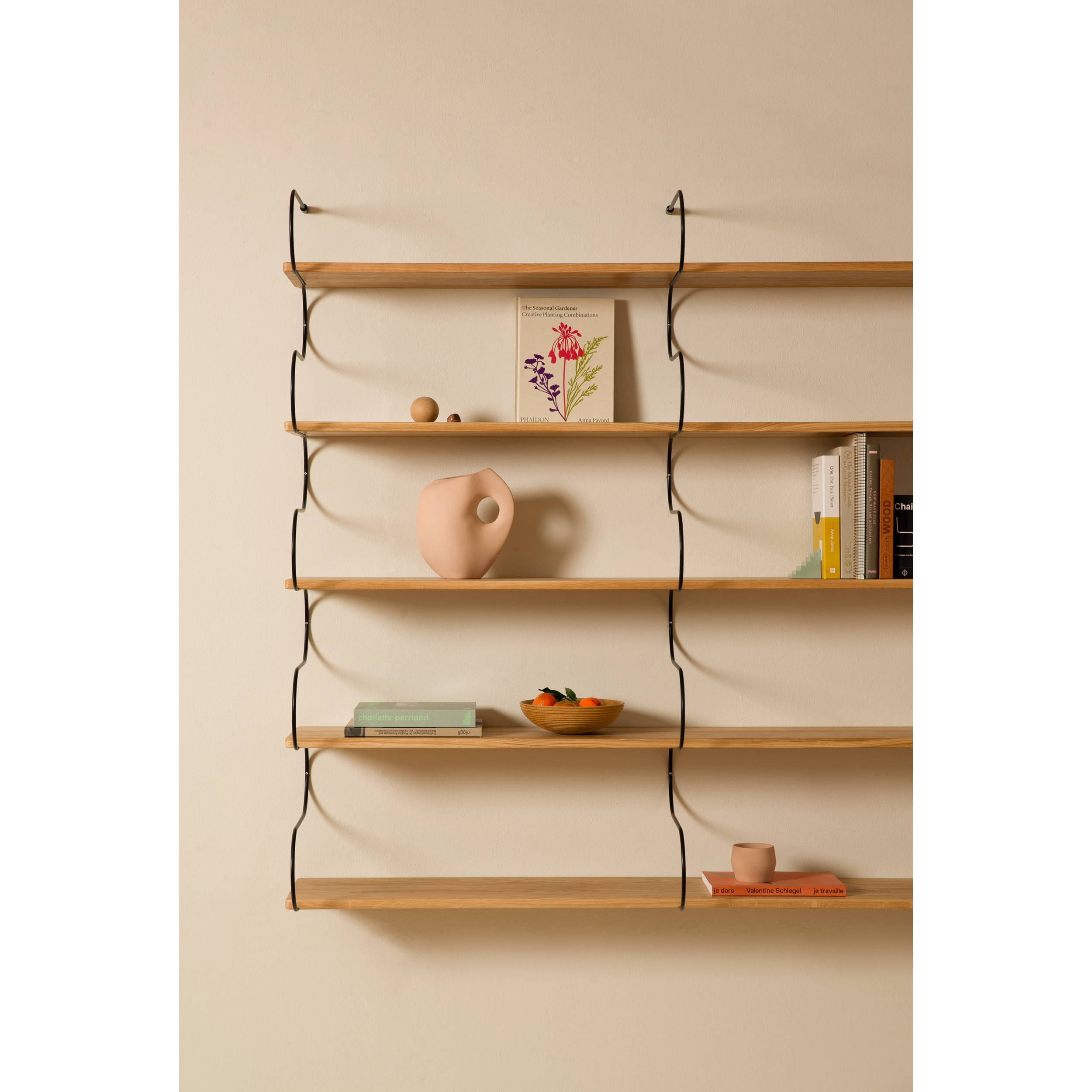 The Onda Double shelves by Schneid, made from oak, feature wooden planks and black brackets that elegantly display a stylized vase, books, wooden spheres, and a fruit bowl against a beige backdrop.