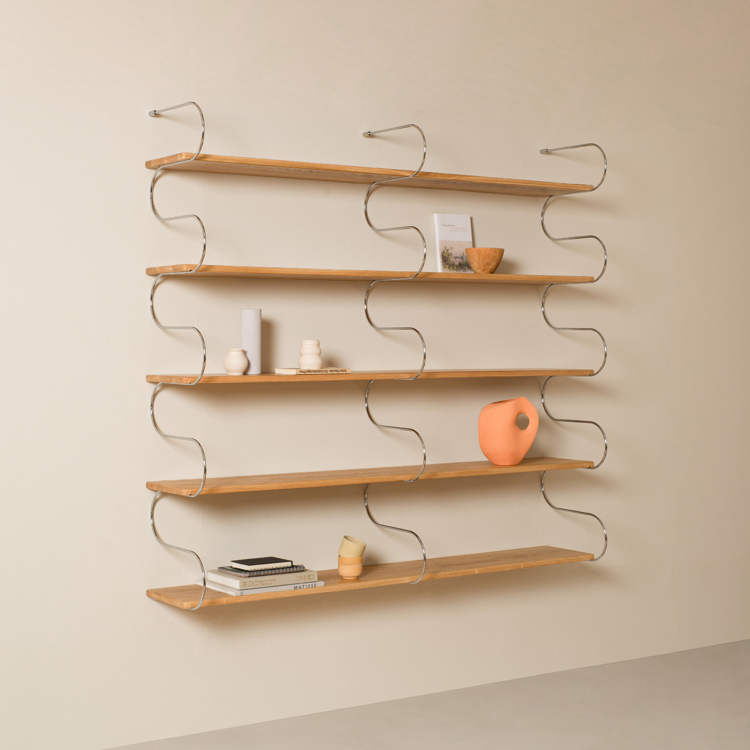 The Onda Double by Schneid is a minimalist wall-mounted shelf with four oak planks supported by wavy metal brackets, perfect for displaying small items like books, candles, and vases against a neutral wall.