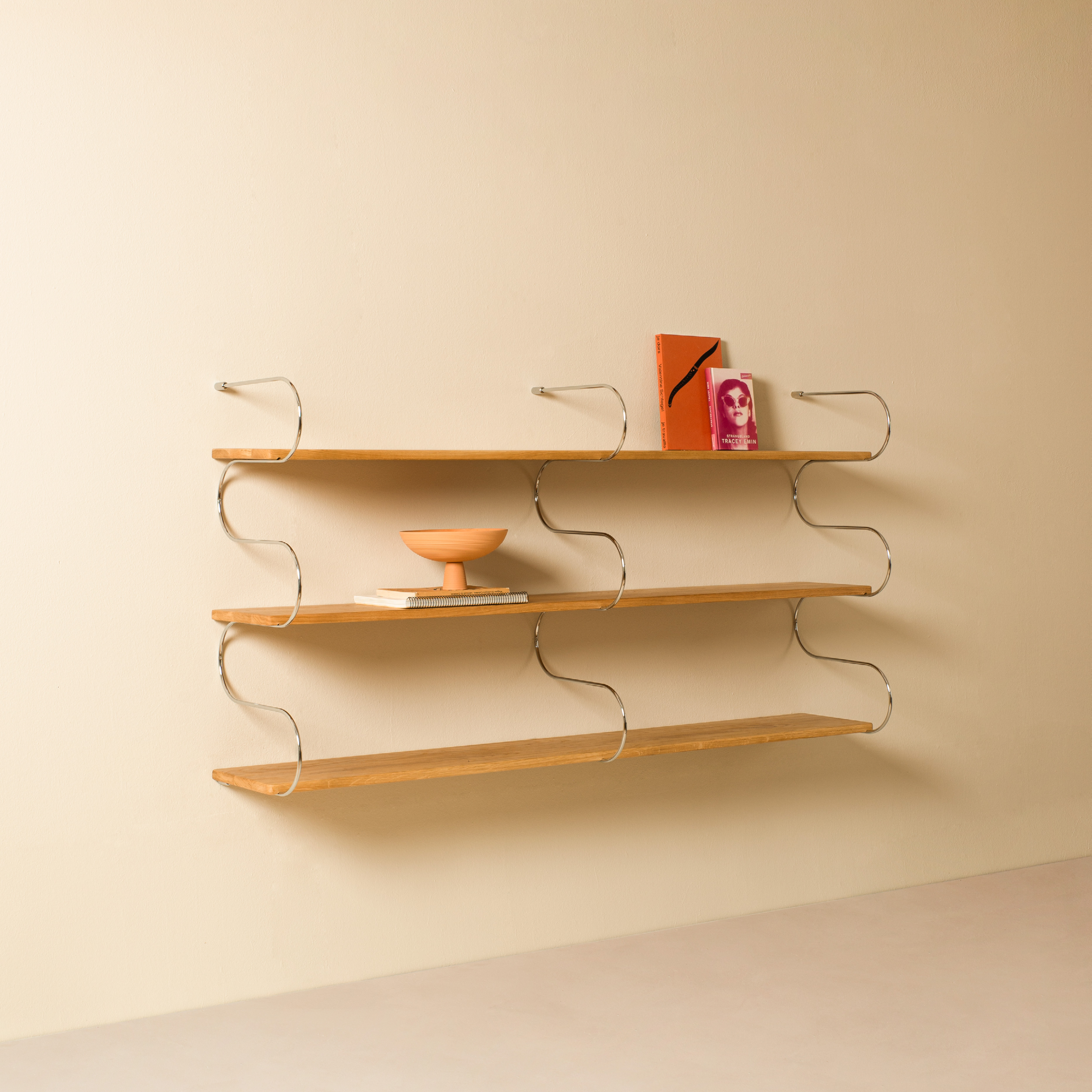 The Onda Double by Schneid features minimalist oak wall shelves with curved metal supports, holding a small bowl and two books. Set against a beige wall with a light floor, it creates a simple, modern aesthetic.