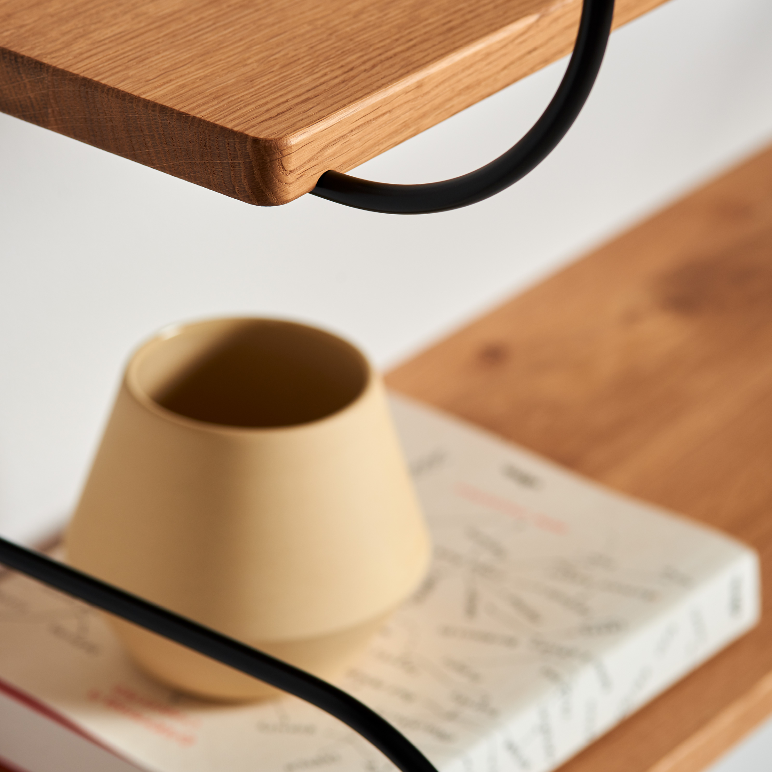 A close-up of an oak wood shelf showcases a beige cup on a book, supported by sleek black metal braces. Part of the Schneid Onda Double modular system, it highlights the shelfs corner against a softly blurred backdrop.
