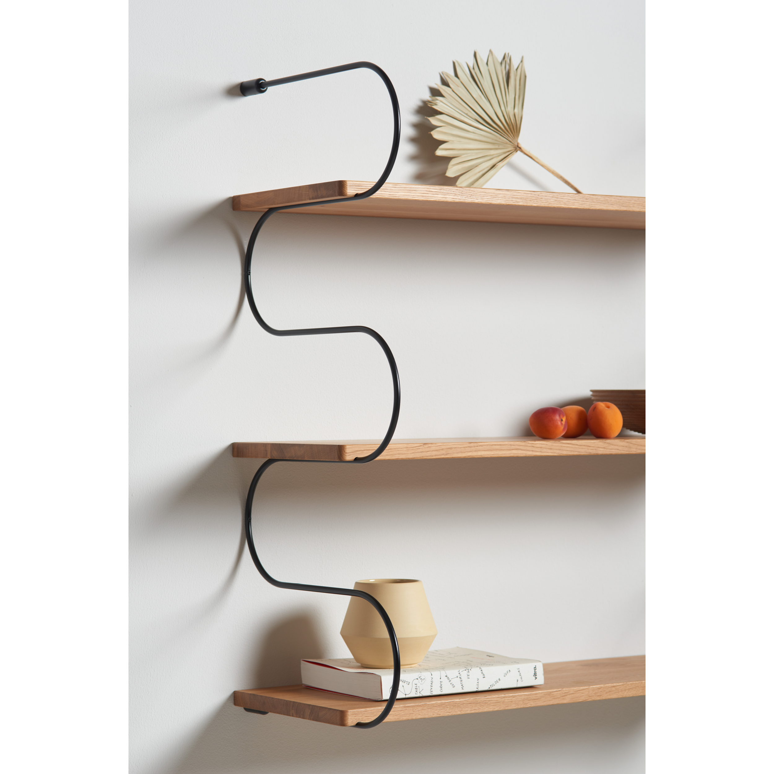 The Onda Double by Schneid is a modular, minimalist shelving system. It features oak wood shelves with wavy black metal supports and displays a dried fan palm leaf, red fruits, a book, and a beige vase, showcasing an emphasis on natural textures.