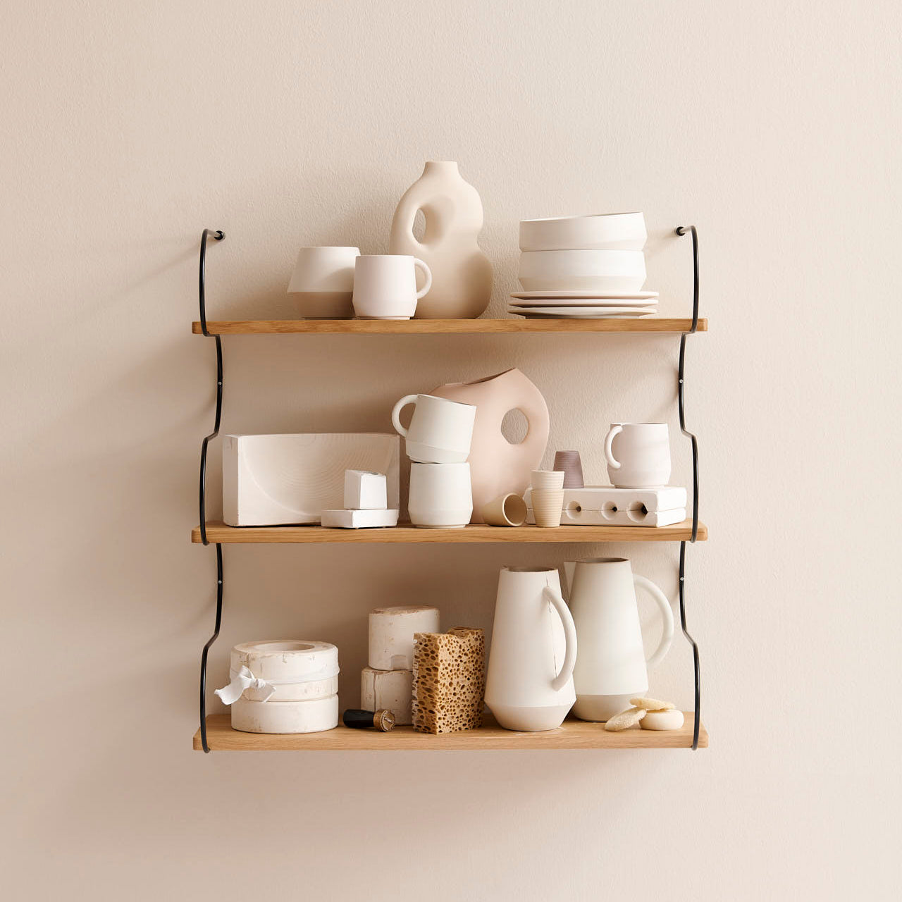 The Onda shelf by Schneid, crafted from oak wood and mounted on a beige wall, beautifully displays minimalist cream-colored pottery including mugs, bowls, vases, and plates. The potterys geometric shapes and textured surfaces add a modern and artistic touch to the arrangement.