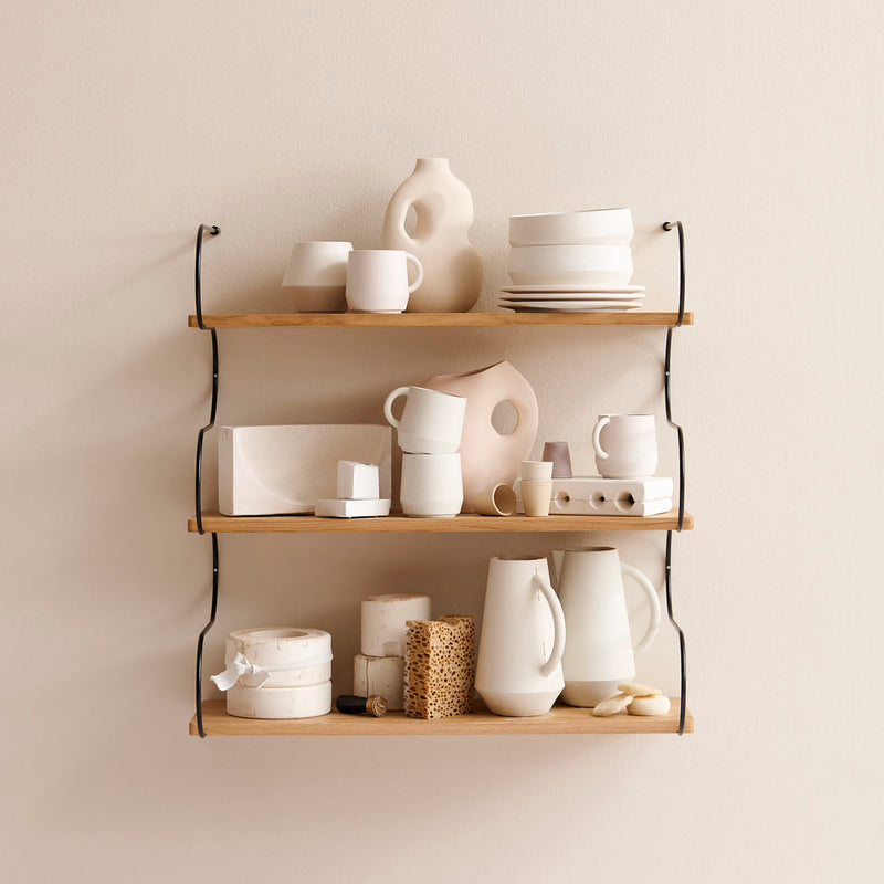 The Onda shelf by Schneid, crafted from oak wood and mounted on a beige wall, beautifully displays minimalist cream-colored pottery including mugs, bowls, vases, and plates. The potterys geometric shapes and textured surfaces add a modern and artistic touch to the arrangement.