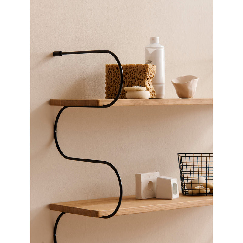 The Onda shelving system by Schneid stands out against a light wall with its oak wood shelves. The top shelf holds sponges, a soap bottle, and soaps, while the bottom supports a wire basket and rectangular objects. Black metal side supports offer modern elegance with subtle curved lines.