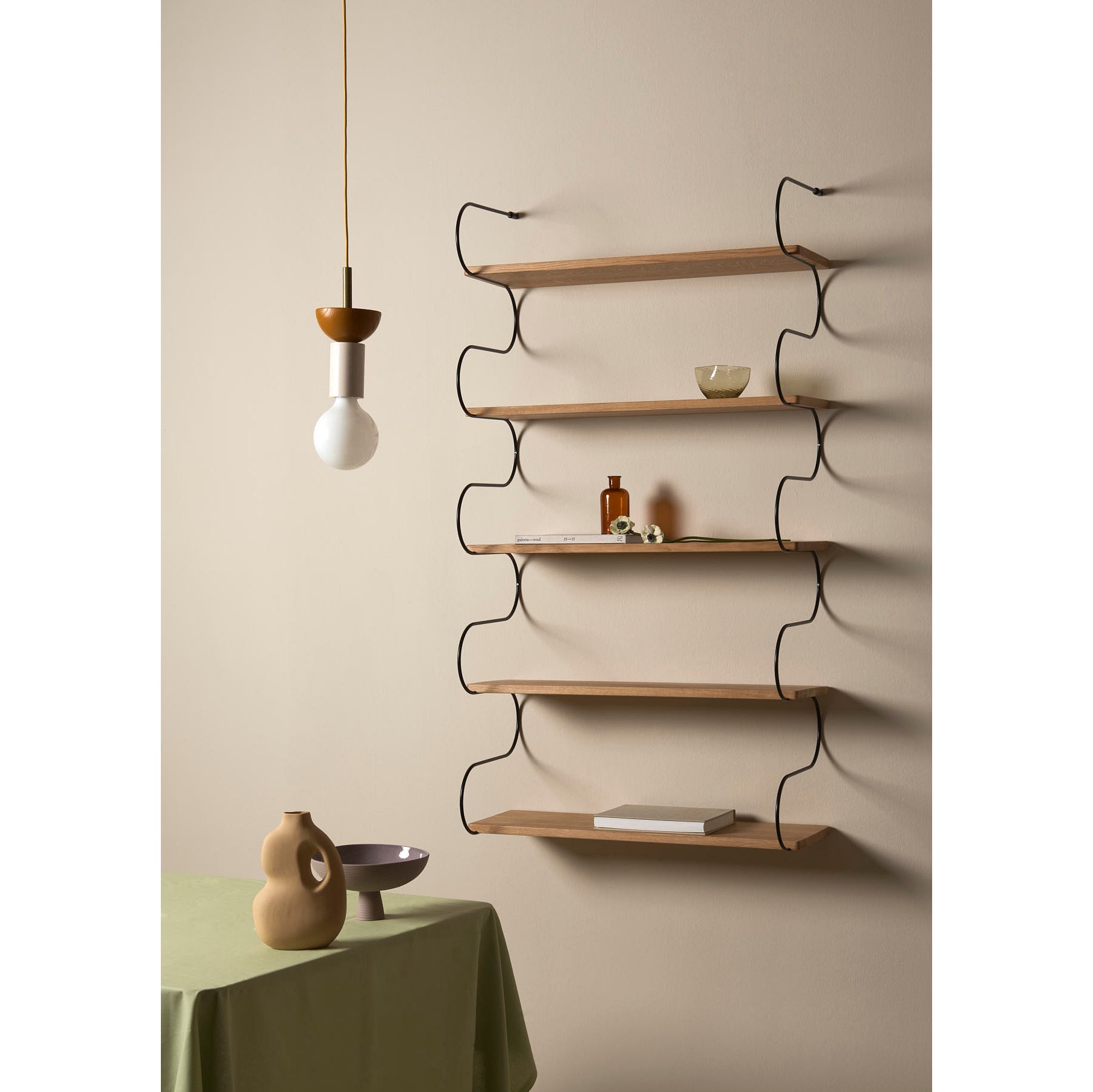 A minimalistic room features the Onda, a wavy oak shelf by Schneid, holding a bowl and small objects. A table with a green cloth hosts two ceramic vases, while above, a modern pendant light casts a soft glow. The decor is defined by simple and elegant curved lines.