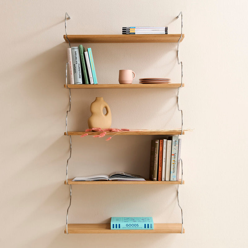 The Onda by Schneid is a wall-mounted modular shelving system featuring five oak shelves, perfect for displaying books, a pink cup, a beige vase, and decorative items like dried flowers. Its thin metal brackets provide support against a light-colored wall.