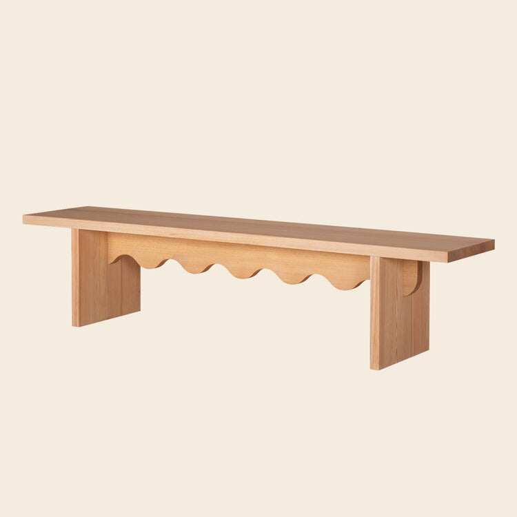 The Tami Bench from Schneid is crafted from light-colored larch wood and features a minimalist design with a smooth top and wavy cut-out support panel, elegantly standing against an off-white background as part of the Tami Dining Collection.