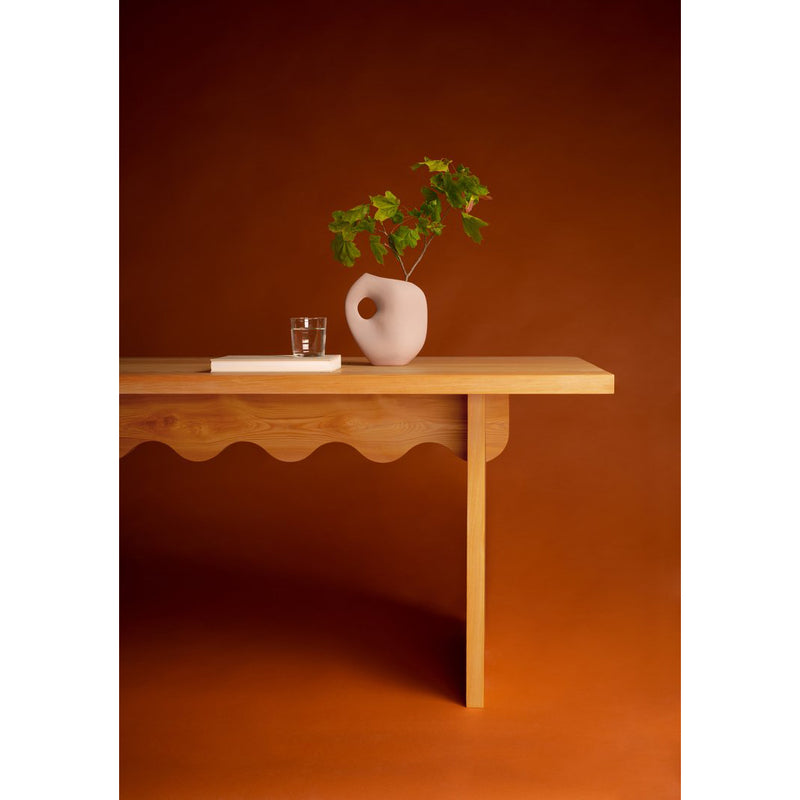 A minimalist Tami Table by Schneid, crafted from naturally oiled larch wood with a wavy edge, holds a white vase with green branches and a glass of water. The warm burnt orange backdrop enhances the earthy elegance of this piece from the Julia Jessen collection.