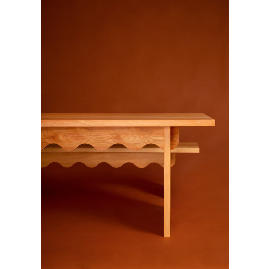 The Tami Table by Schneid from the Tami Dining Collection showcases a distinctive wavy pattern on a brown background. Made of naturally oiled larch wood, it features a smooth tabletop with clean lines and a lower shelf mirroring its shape.