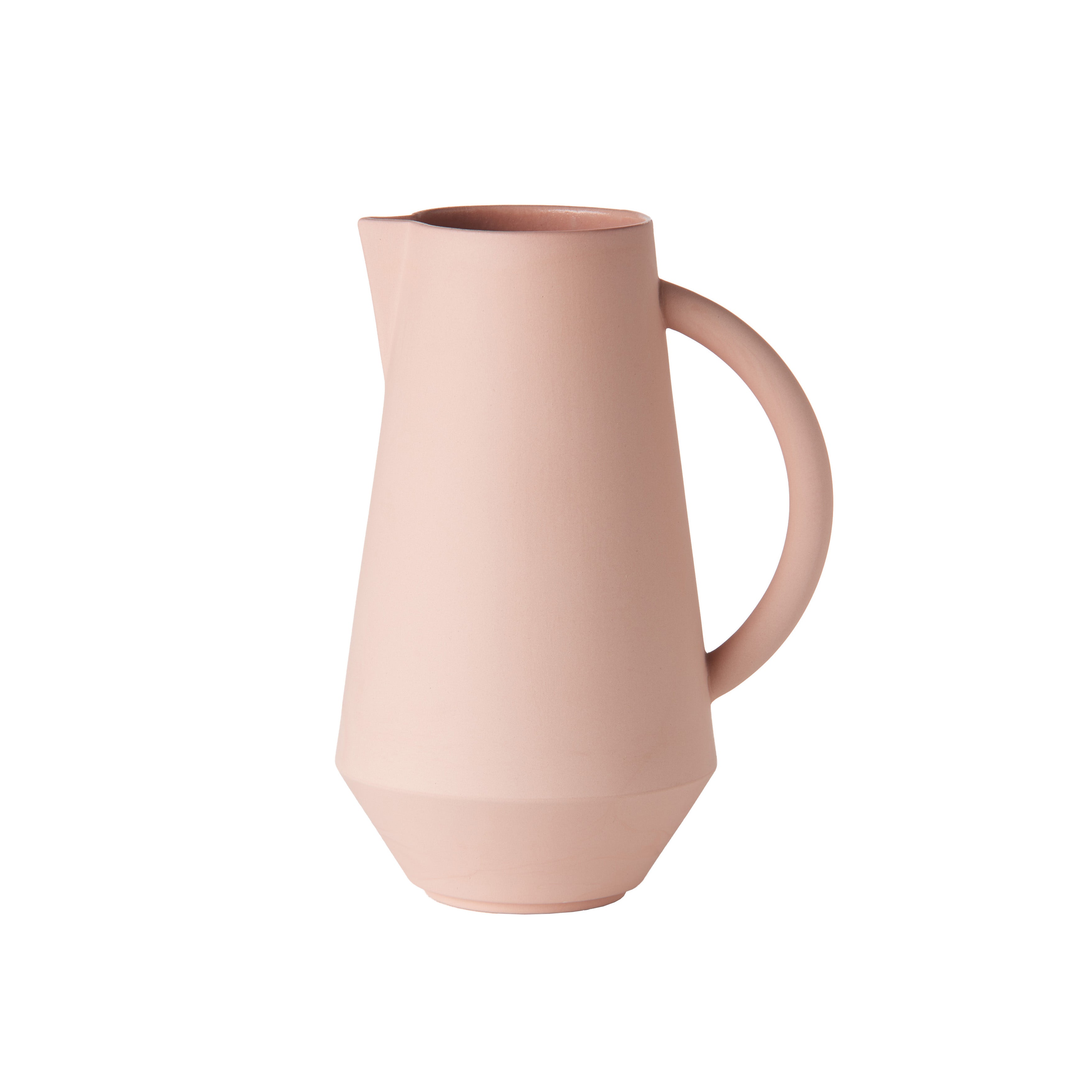 The Schneid Unison Carafe is a minimalist, matte pastel pink ceramic pitcher with a round handle and a narrow spout. Handcrafted in Germany, it features a geometric, angular base for modern elegance, complementing other ceramic tableware against plain white backgrounds.