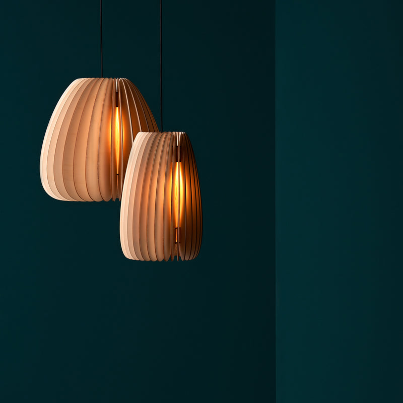Two modern Secundum pendant lights by Schneid, with vertical ribbing, hang from the ceiling. They emit a naturally warm light against a dark teal background. The poplar wood texture contrasts elegantly with the deep-colored wall.