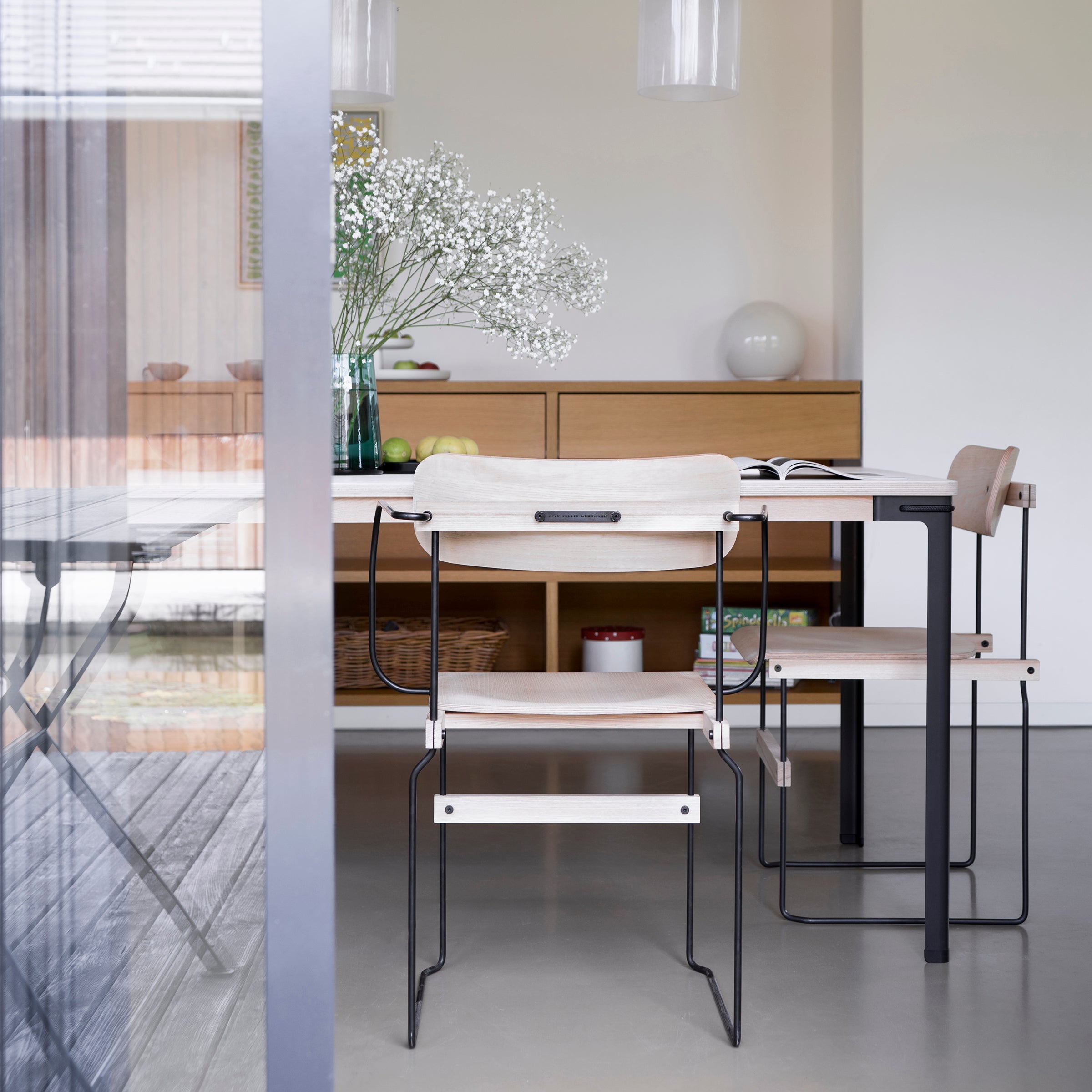A modern dining room showcases a minimalist wooden table paired with Moormann Bruto chairs, featuring light hues and black metal frames. Decor includes a vase with white flowers, an open book, and wooden shelves. Glass doors reflect the setting, emphasizing the elegant ash veneer accents.