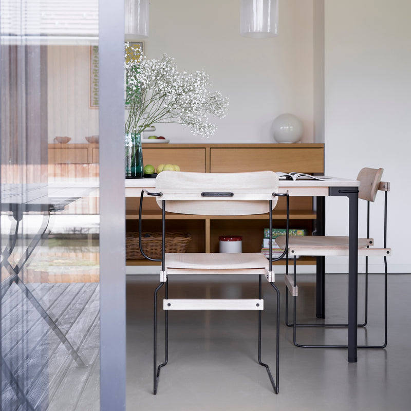 A modern dining room showcases a minimalist wooden table paired with Moormann Bruto chairs, featuring light hues and black metal frames. Decor includes a vase with white flowers, an open book, and wooden shelves. Glass doors reflect the setting, emphasizing the elegant ash veneer accents.