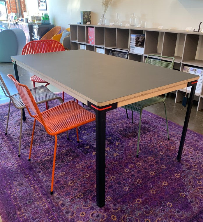 A stylish Moormann Seiltänzer Table (on sale) with a gray tabletop and black steel legs, surrounded by vibrant red, orange, and green chairs on a purple-patterned rug. The well-lit room features shelves and assorted decor.