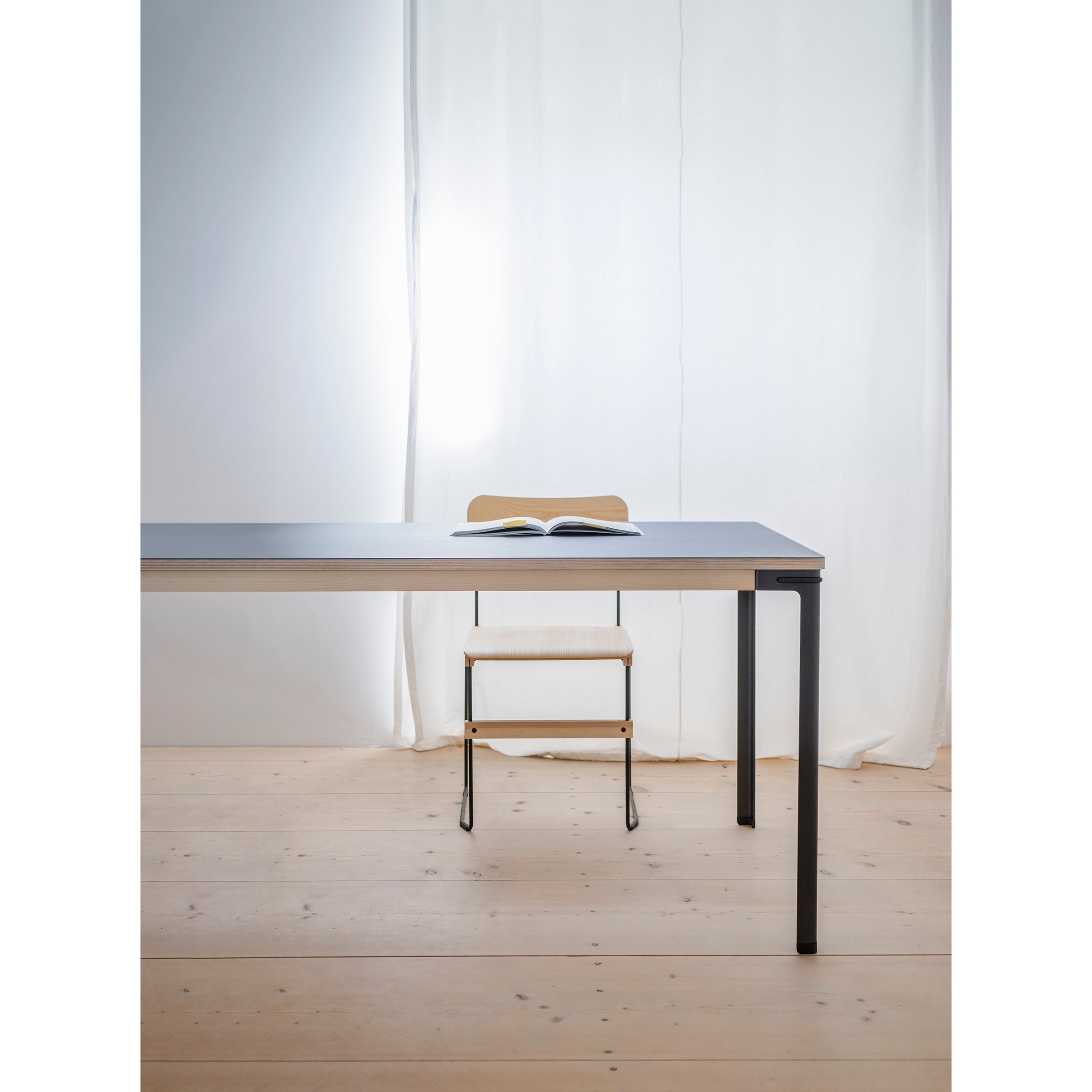 A minimalist room features a sleek table with black legs on a wooden floor. An open book sits at the center, accompanied by a Moormann Bruto chair beneath. Soft light filters through a white curtain in the background.