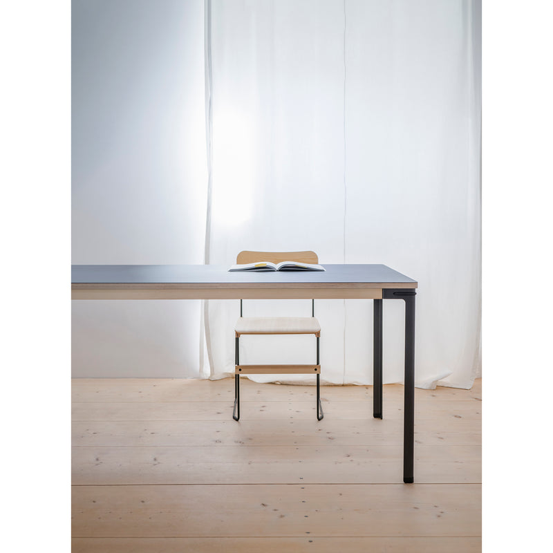A minimalist room features a sleek table with black legs on a wooden floor. An open book sits at the center, accompanied by a Moormann Bruto chair beneath. Soft light filters through a white curtain in the background.