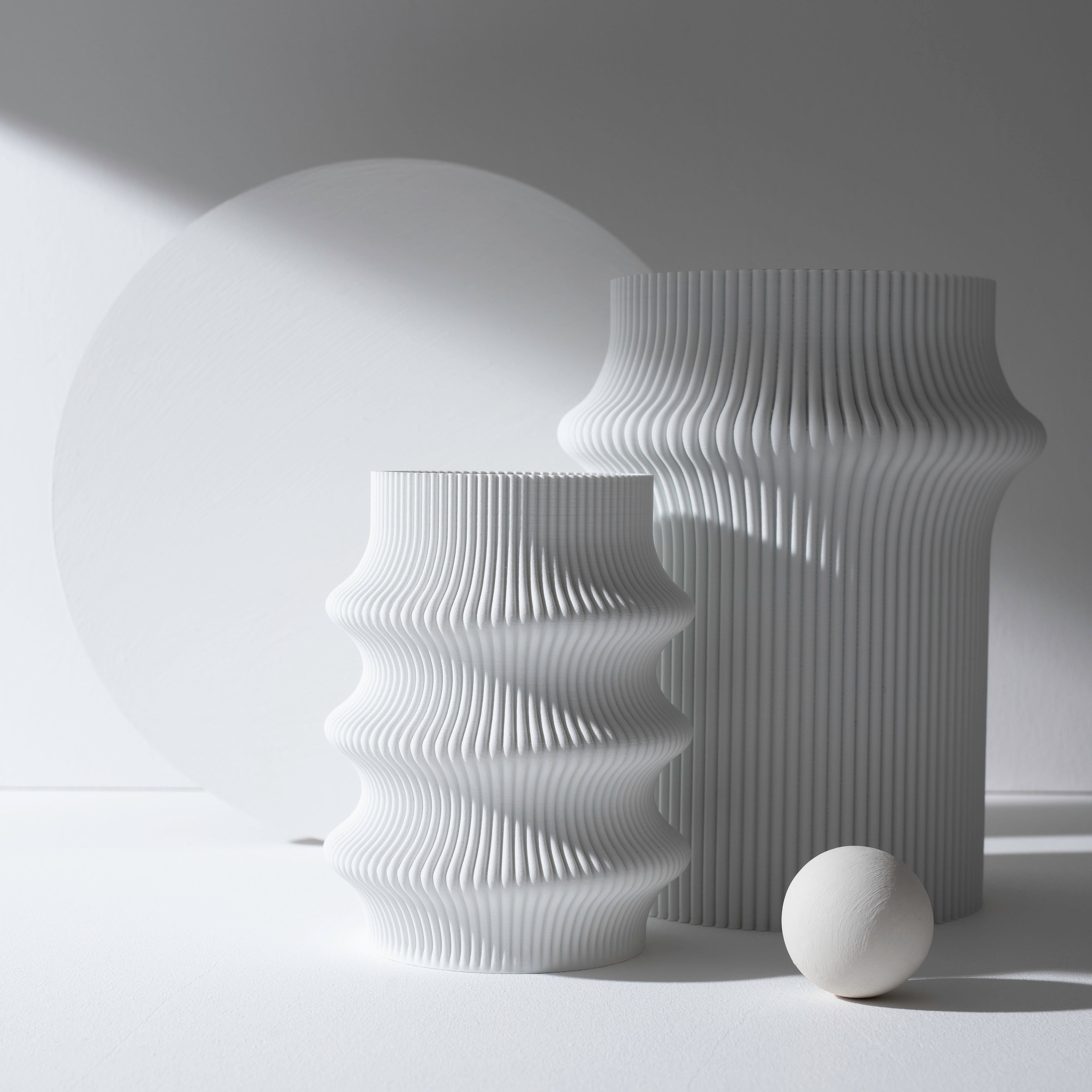 The minimalist still life features two white Zayl 247g vases by Sheyn, resembling architectural pillars, plus a white ball and a round backdrop. The eco-friendly setup is softly lit with natural light, creating gentle shadows.