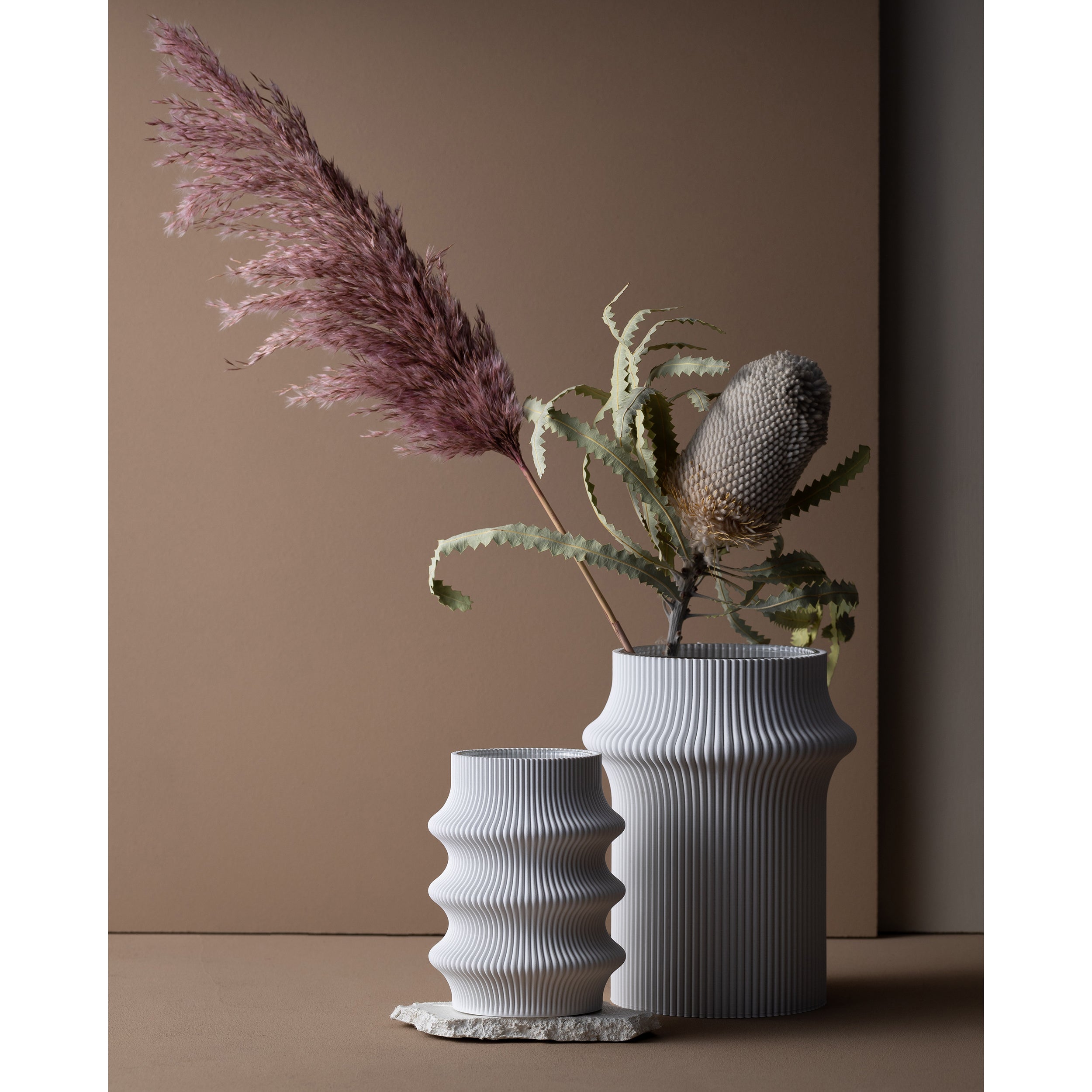 The Zayl 247g Vase by Sheyn, a white ribbed ceramic piece, features a modern, wavy design on a table against a beige background. Made from eco-friendly materials, the smaller vase holds dried pampas grass, while the larger presents assorted dried foliage.