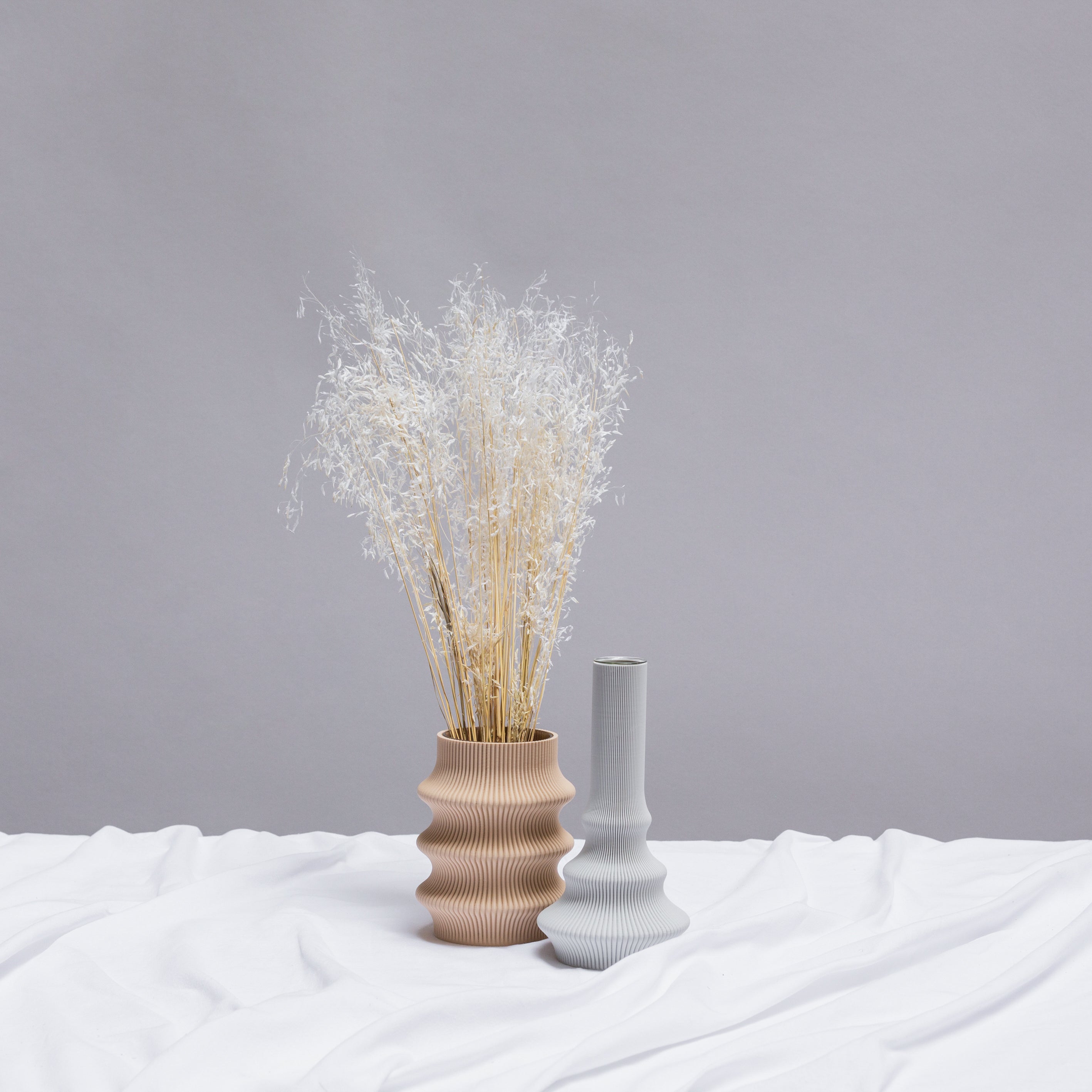 The scene features two vases on a white cloth: the larger wavy beige one, crafted via 3D printing and holding feathery dried grasses, alongside a smaller similarly textured gray vase. Set against a plain gray backdrop, these elegant pieces are crafted by Sheyns innovative design studio, including their Zayl 114g Vase.
